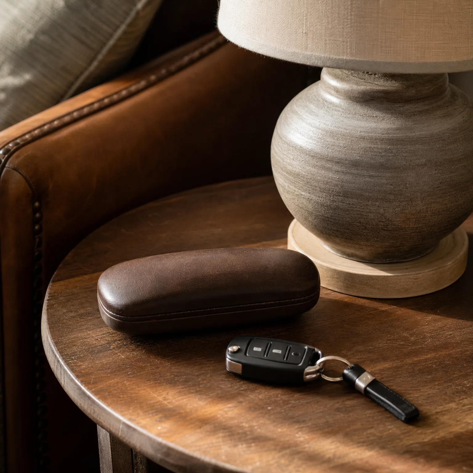 A brown hard-shell eyeglass case and a black folding car key with a leather fob rest on a round wooden side table beside a ceramic lamp base, with a brown leather armchair behind.