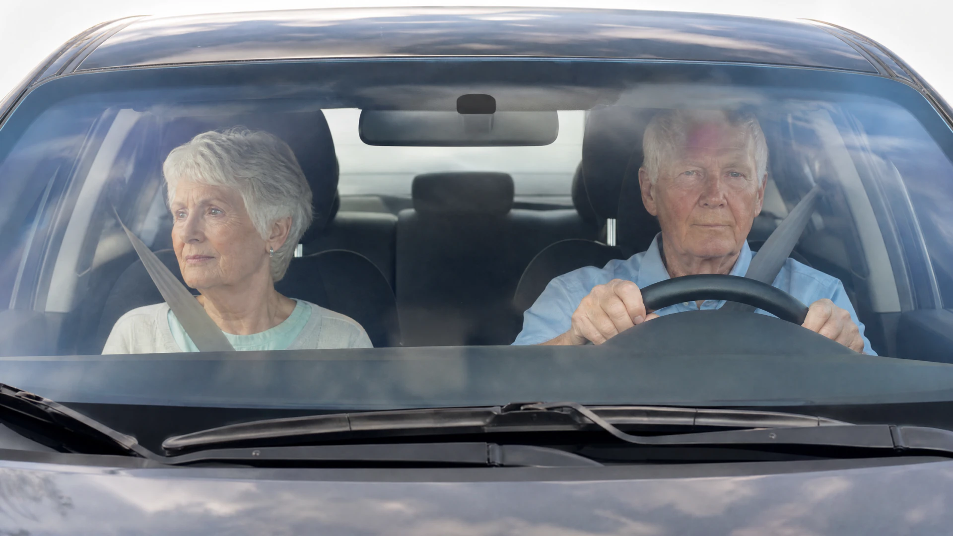 An elderly man and woman sit buckled in the front seats of a car, seen through the windshield.