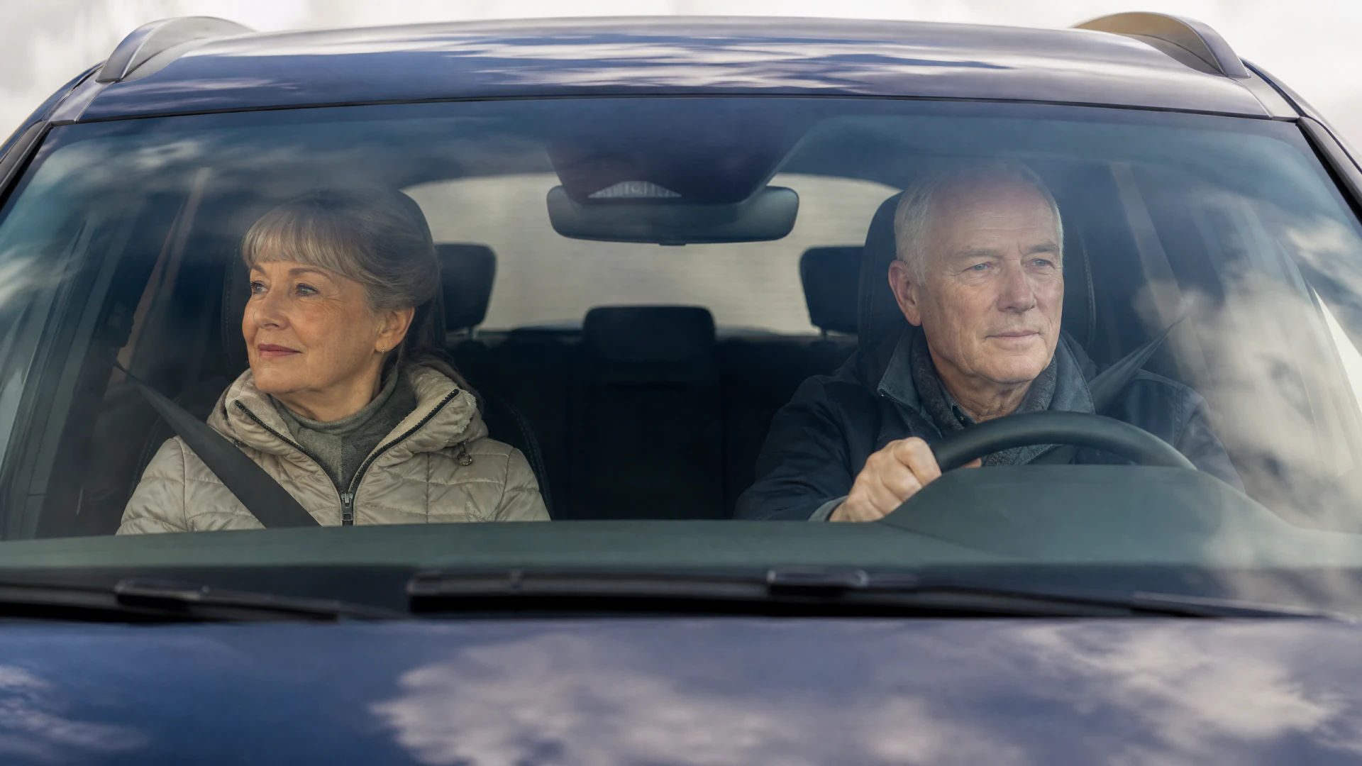 An older man and woman sit in the front seats of a dark blue car, viewed through the windshield.