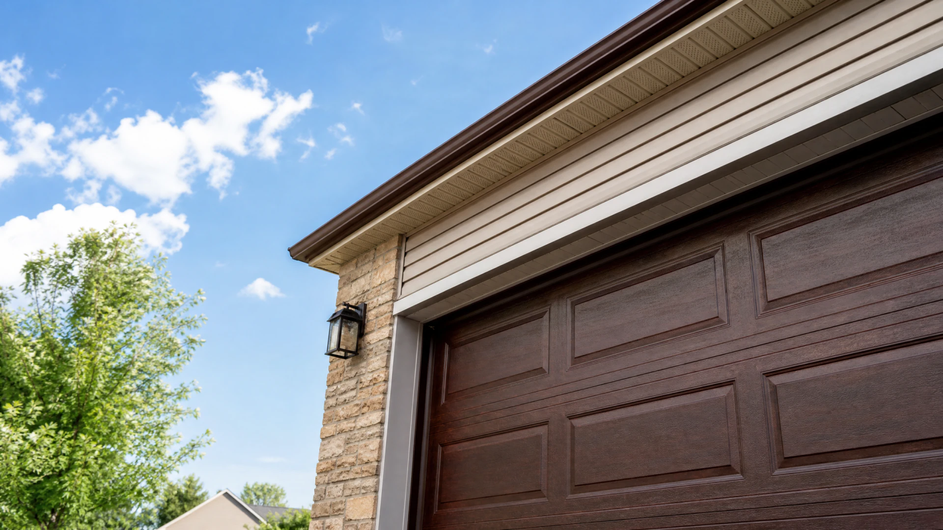 Low-angle exterior view of a residential garage with a dark brown raised-panel door, natural stone facade, and a black lantern wall light beneath a beige vinyl soffit.