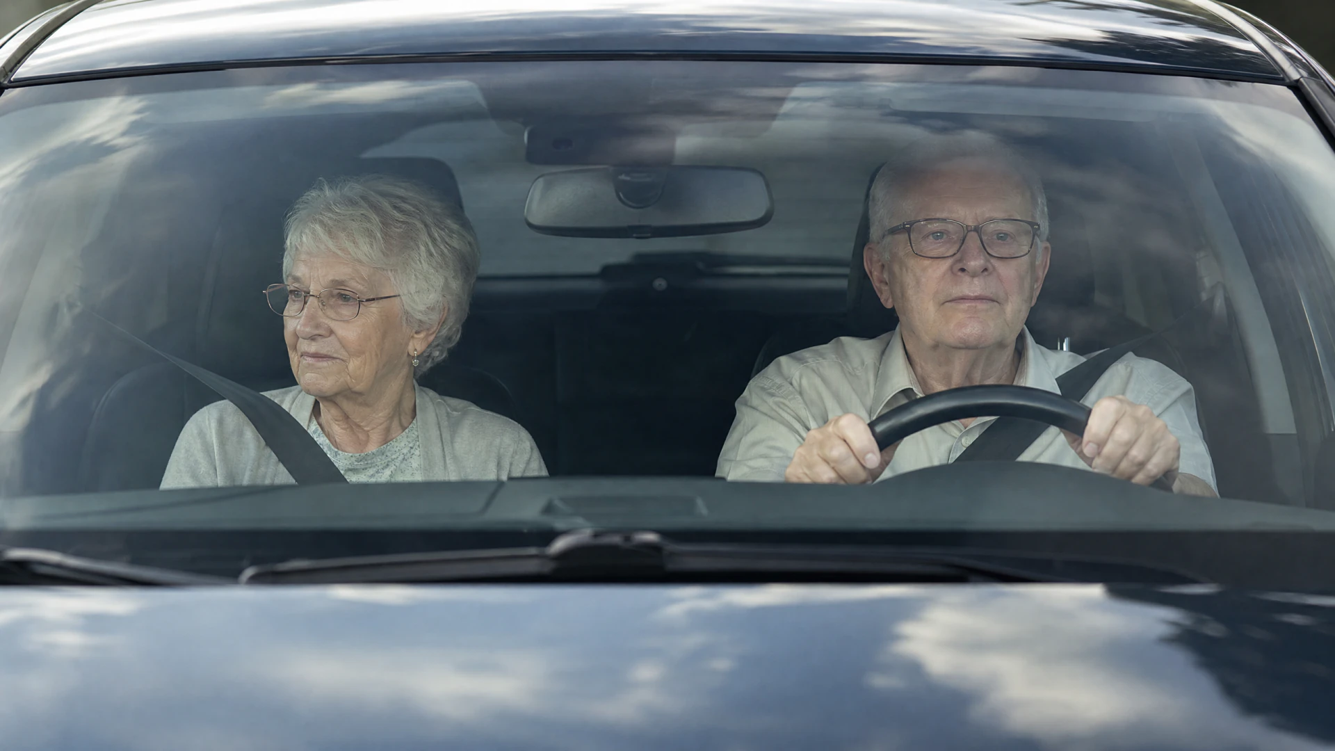 An elderly couple sit in the front seats of a car, viewed through the windshield, with the man driving and both wearing seatbelts and glasses.