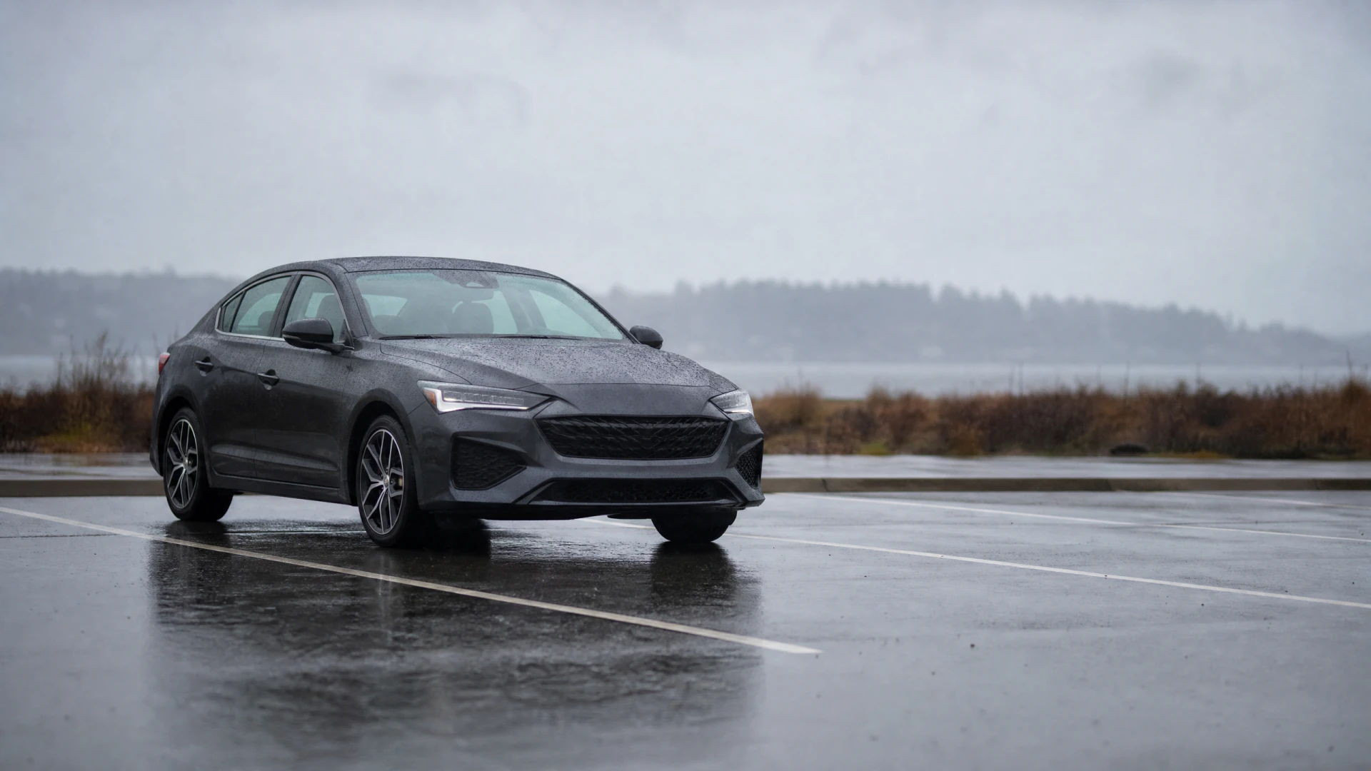 A wet dark gray fastback sedan parked in an empty rain-soaked parking lot under an overcast sky.