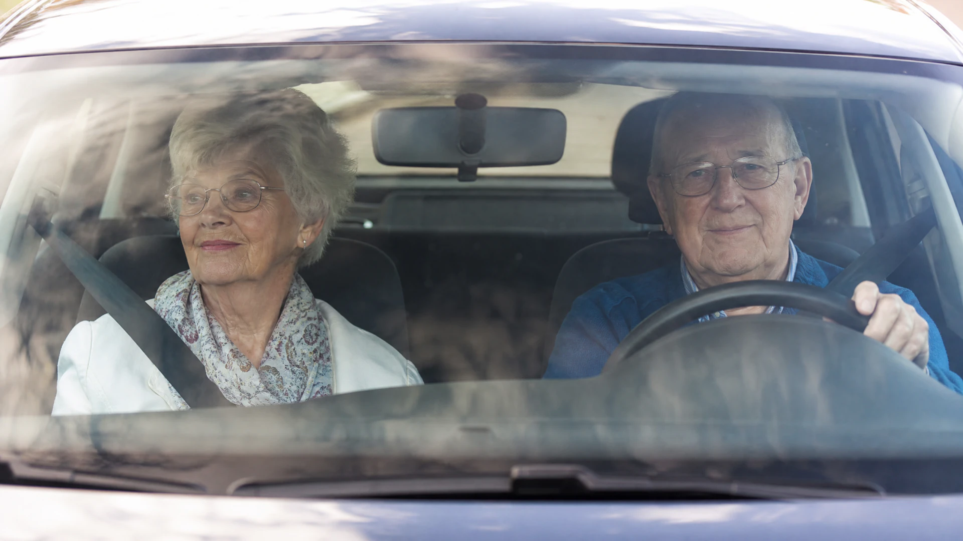 An elderly man and woman sit in the front seats of a car, photographed through the windshield.