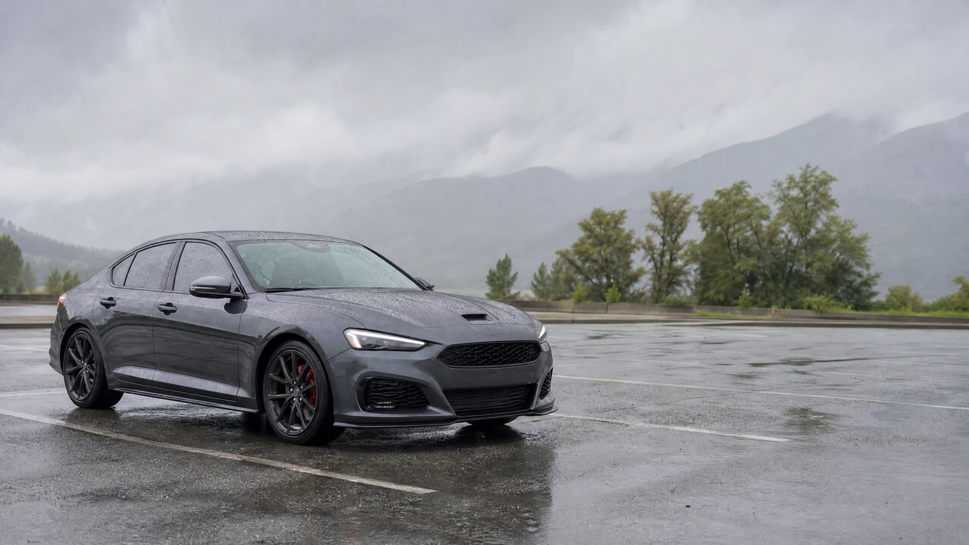 A dark gray sport sedan with a hood scoop and red brake calipers parked on a rain-soaked lot with misty mountains in the background.