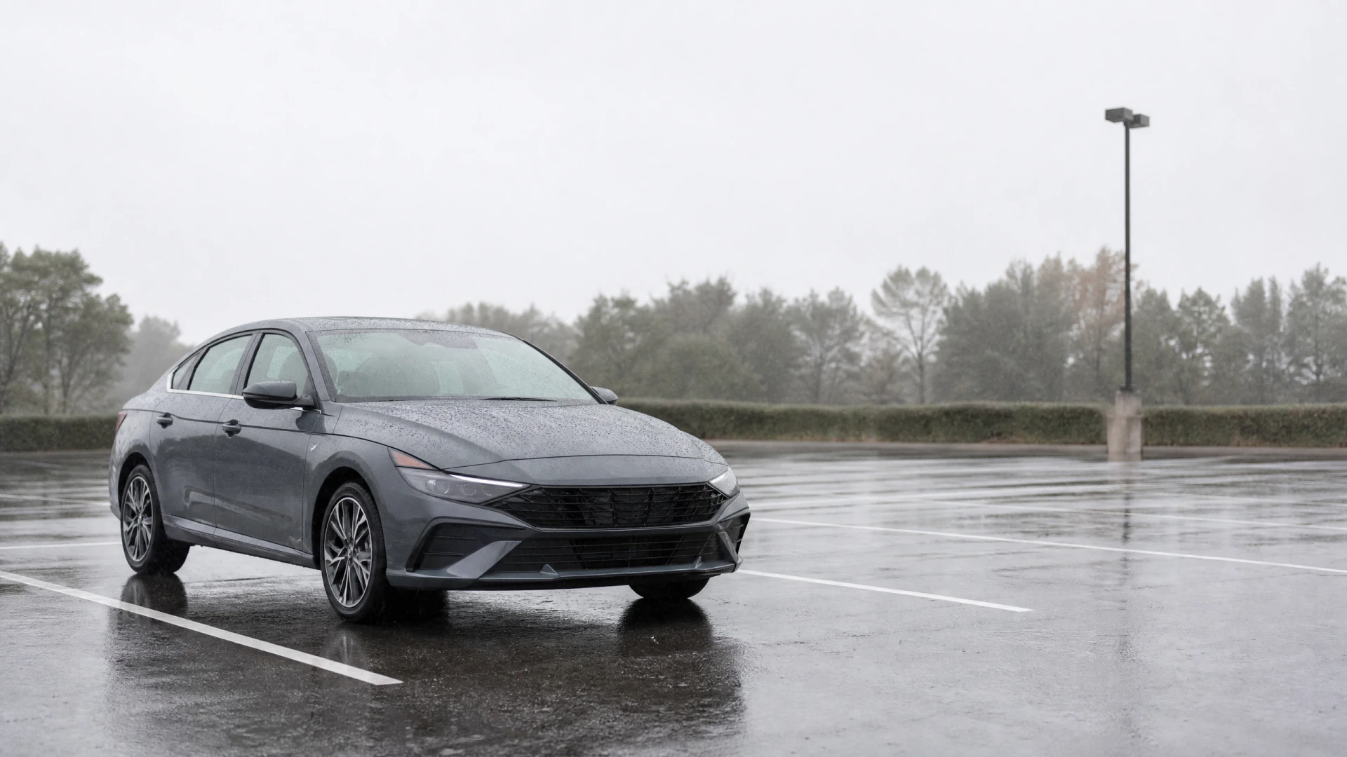 A wet gray sedan sits alone in an empty parking lot on a rainy, foggy day.