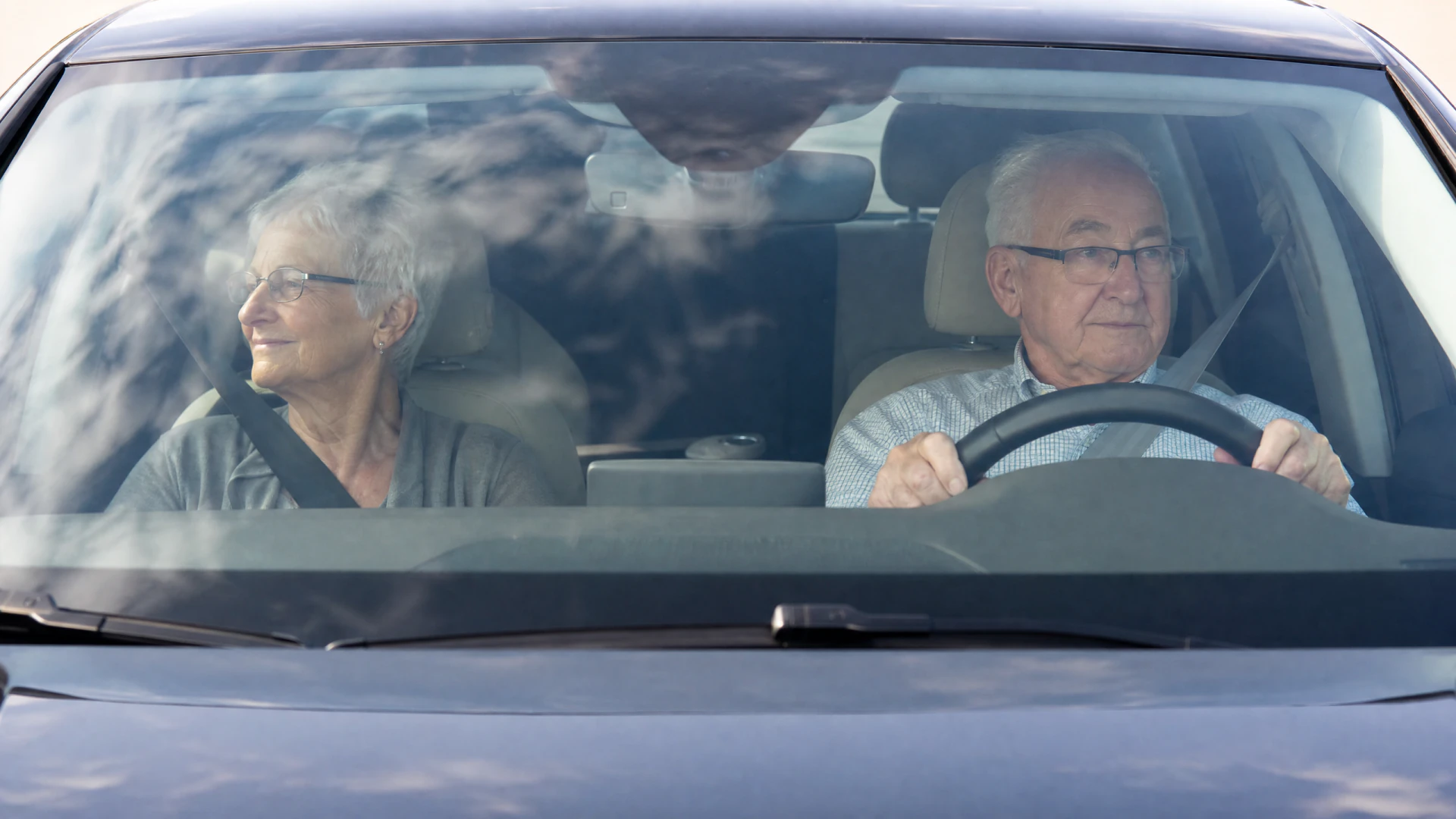 An elderly man and woman sit in the front seats of a car, both wearing seatbelts, as the man drives.