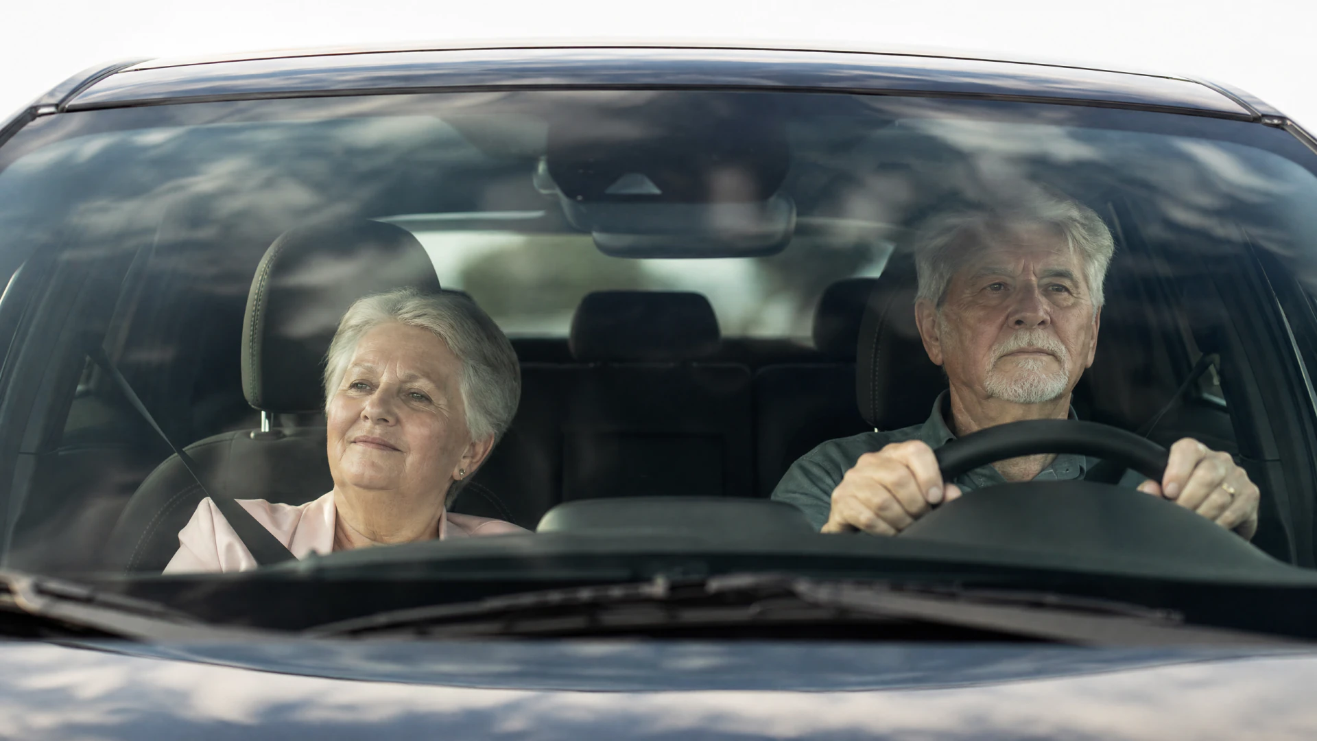 An elderly couple sits in the front seats of a car, viewed from outside through the windshield, with the man driving and the woman in the passenger seat.