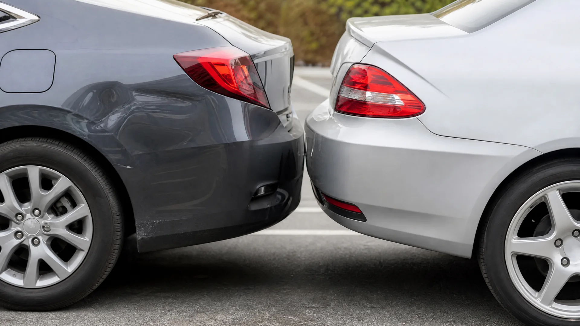 Rear bumpers of a dark gray car and a silver car touching in a parking lot, with visible dents and scrapes on the panels.