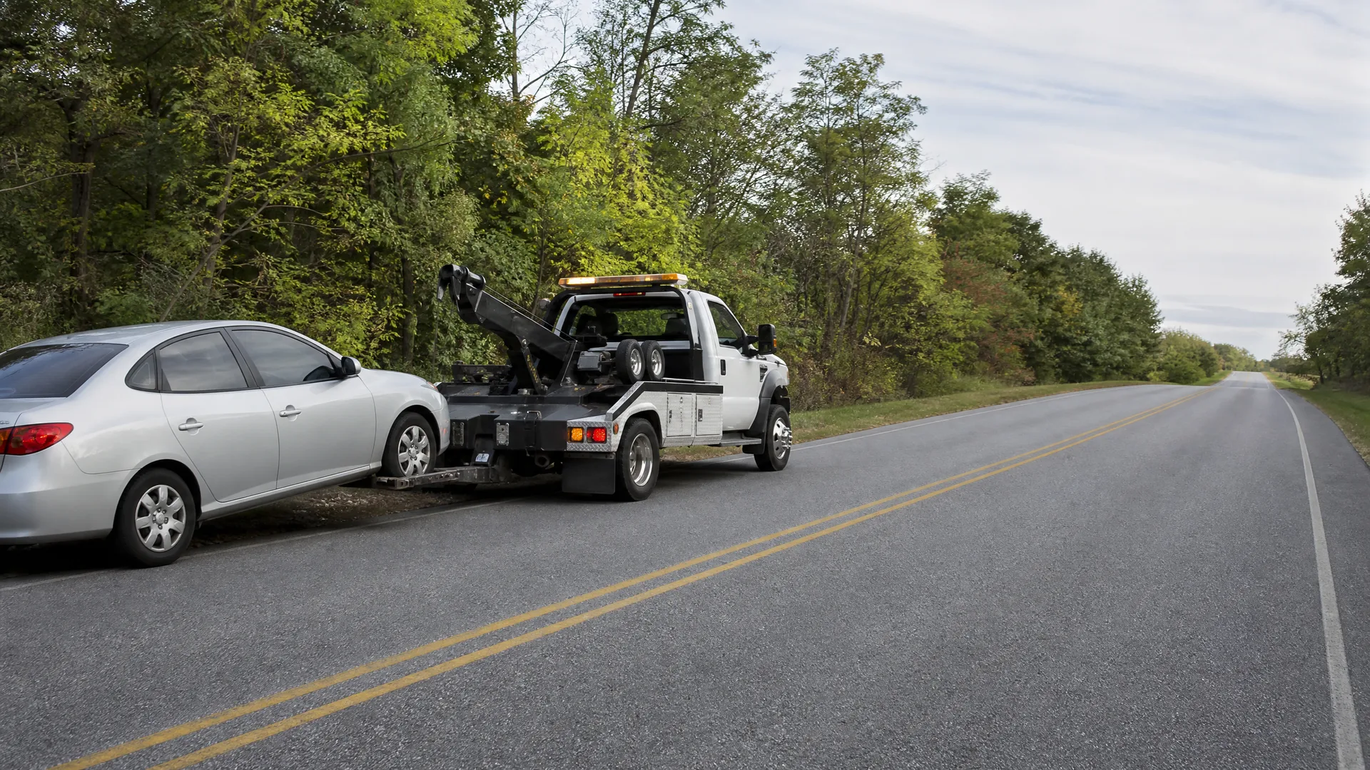 A white tow truck parked on the shoulder of a two-lane road with yellow center lines, hooking up a silver sedan, with dense green trees along the roadside.