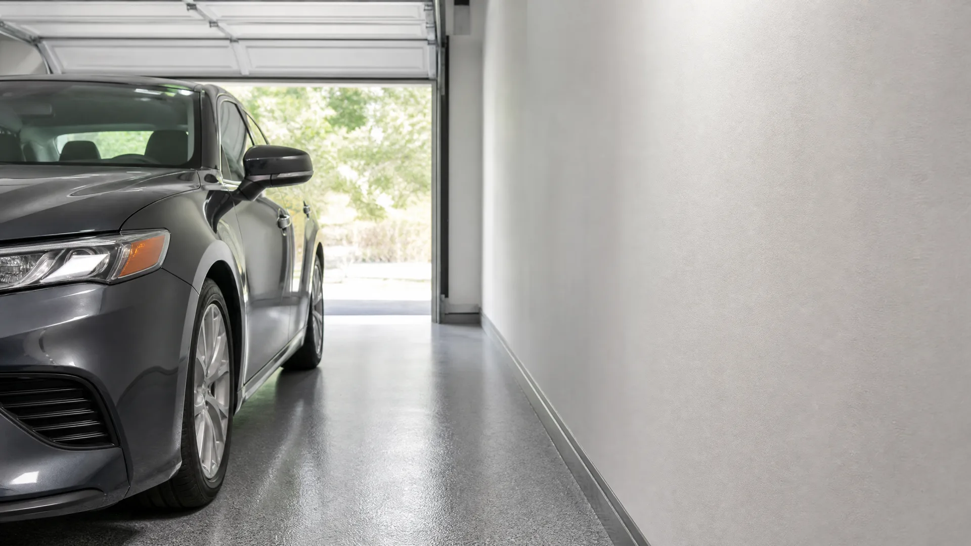 Gray sedan parked inside a garage with a polished speckled floor, a plain light wall on the right, and an open garage door showing green trees outside.