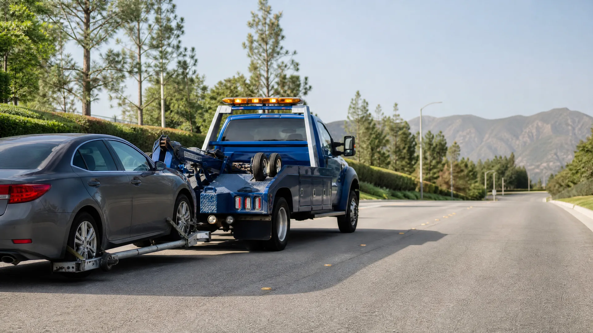 A blue tow truck towing a gray sedan by its front wheels on a paved road, with trees, a streetlight and distant mountains in the background.