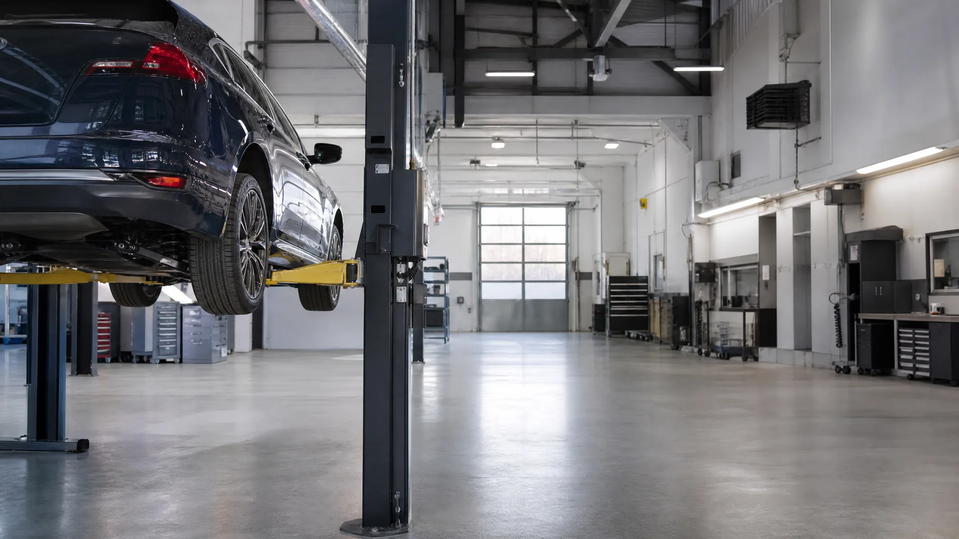 A dark blue SUV raised on a two-post lift inside an empty workshop with polished concrete floor, tool cabinets along the walls and a closed roller door at the far end.
