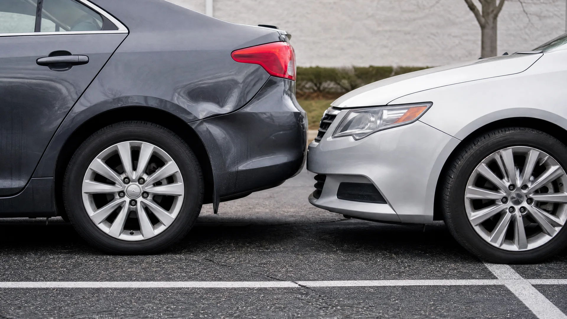 A silver sedan's front bumper touching the dented rear bumper of a dark gray sedan in a paved parking lot, with a light-colored wall behind.