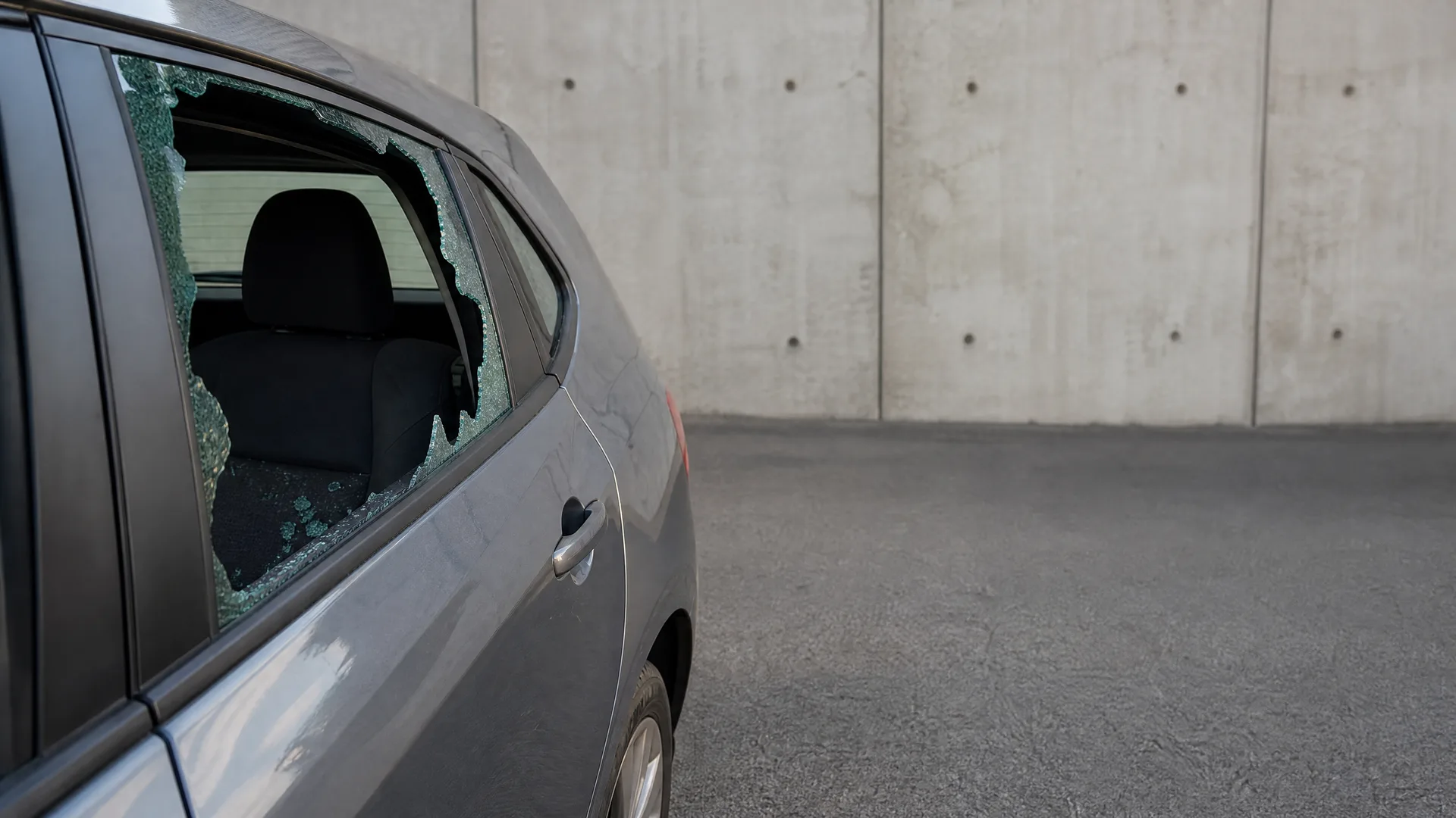 A gray car parked beside a concrete wall with its rear passenger window shattered, leaving jagged green-tinted glass edges and fragments on the sill.