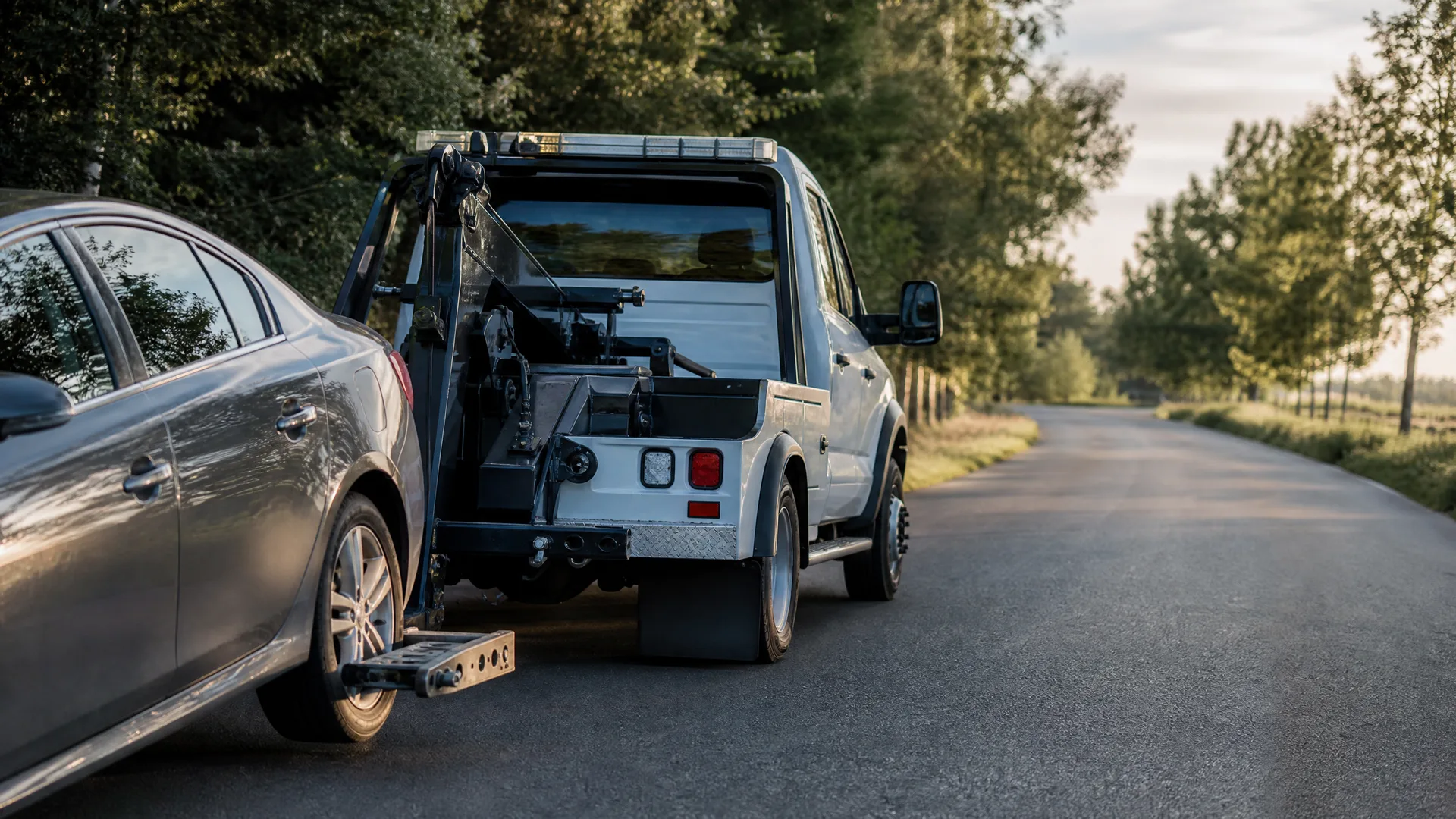 A white tow truck towing a silver sedan by its front wheels on a paved road lined with trees.