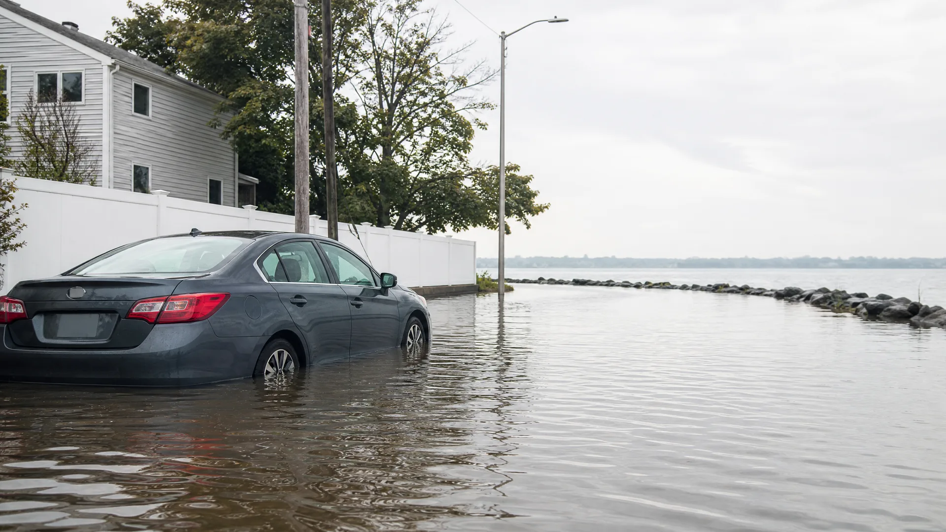 A dark sedan sits in floodwater covering a street beside a white fence and house, with a rock breakwater and open water under an overcast sky.