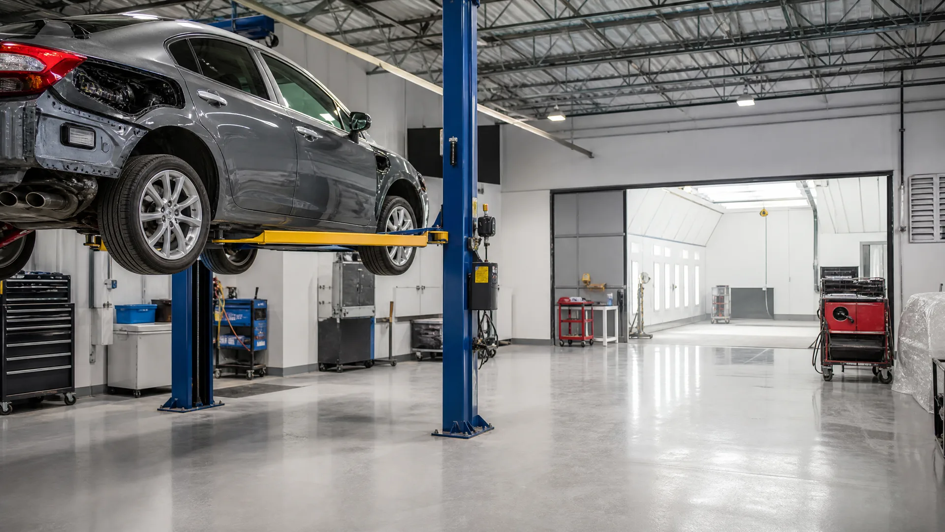 A gray sedan with front-end damage raised on a blue two-post lift inside an auto repair shop with a polished gray floor, tool carts, and an open bay leading to a white paint booth area.