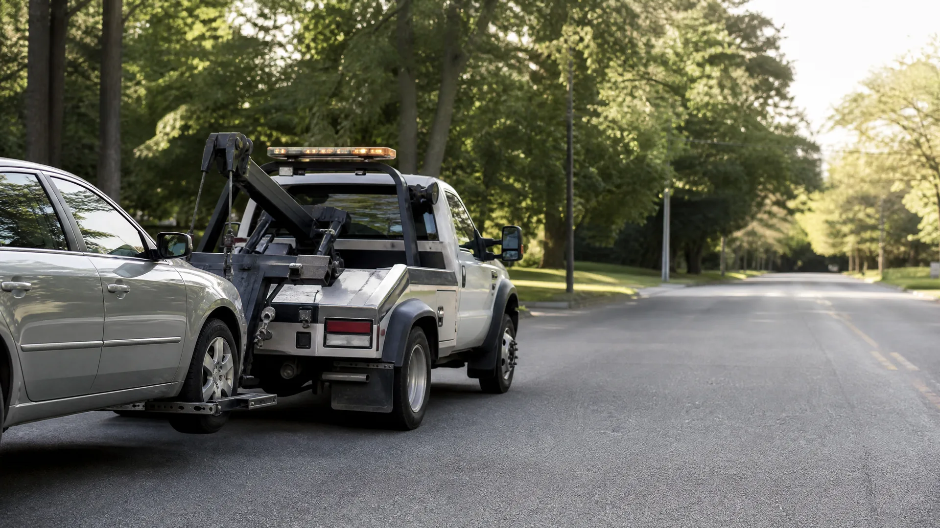 A white tow truck with an amber light bar towing a silver sedan by its front wheels on a paved road lined with green trees.