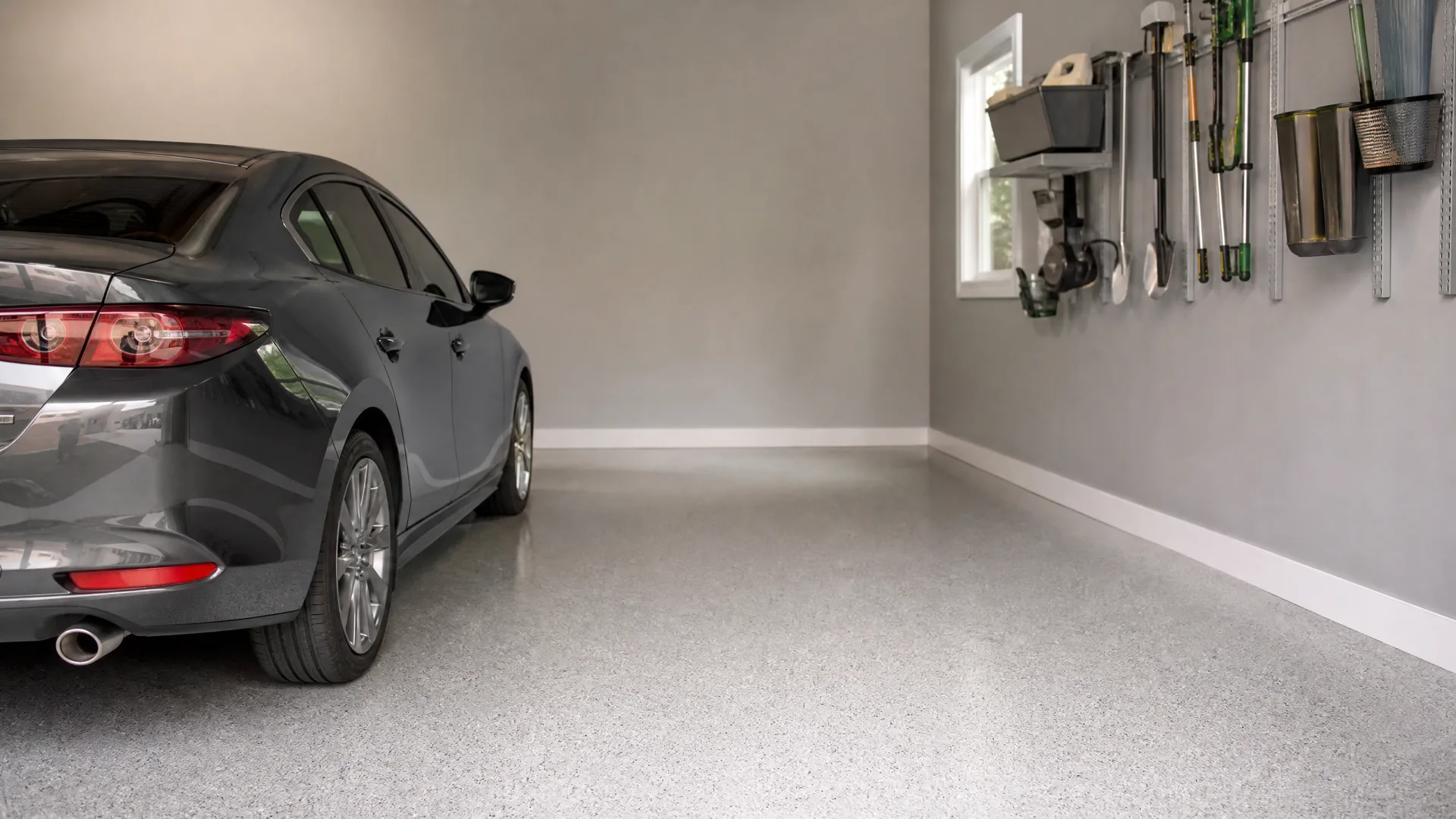 A dark gray sedan parked in a garage with a speckled epoxy-coated floor, gray walls, a small window, and garden tools and bins hung on a wall rail system.