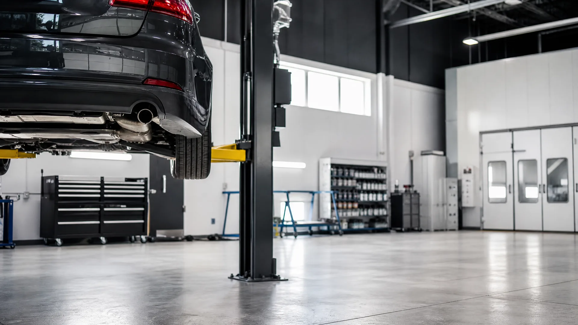 A dark hatchback car raised on a two-post lift inside an automotive shop with a polished concrete floor, tool cabinets, shelved supplies and white paint-booth doors in the background.