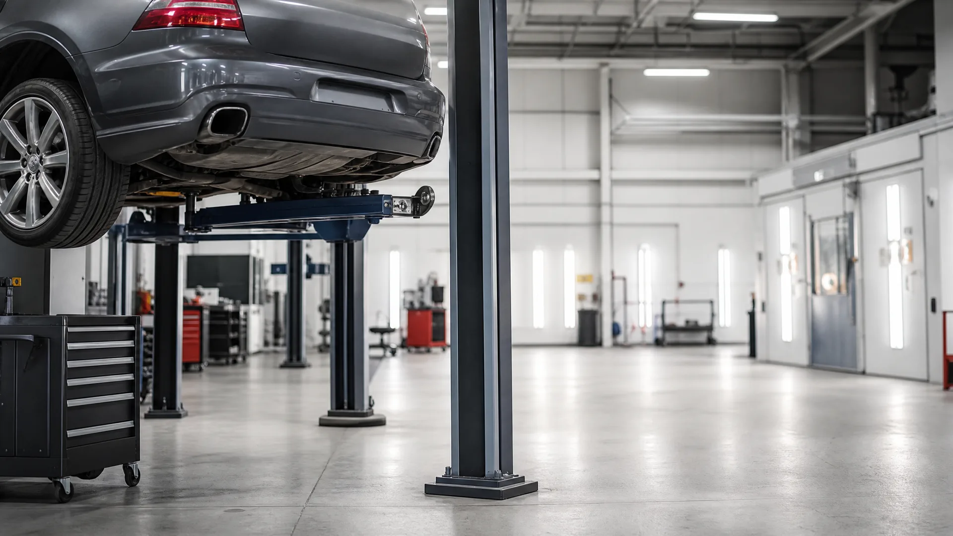 A gray sedan raised on a two-post lift inside a brightly lit garage with polished concrete floor, a rolling tool chest at left and service bays in the background.