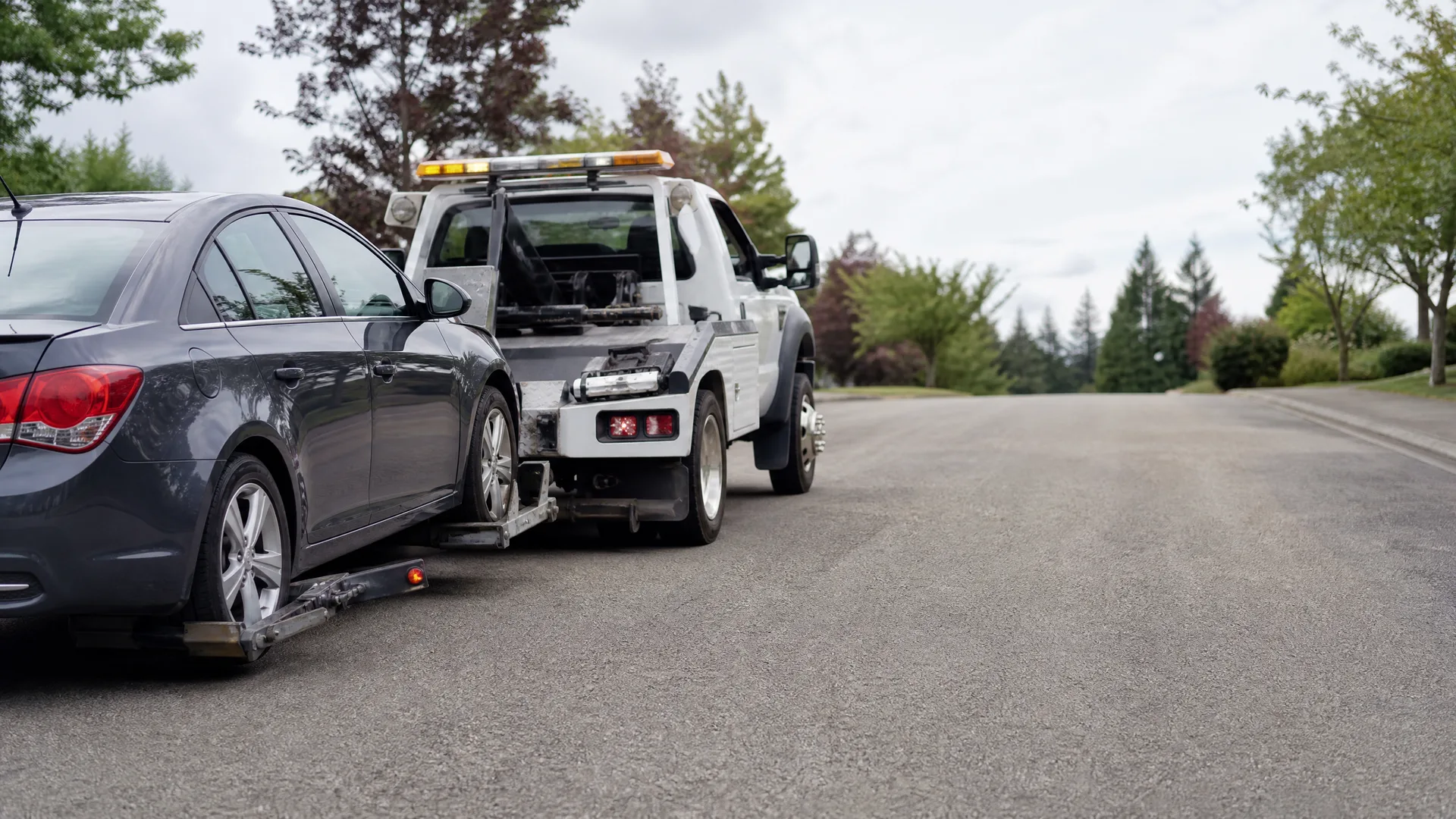 A white tow truck lifts the front wheels of a dark gray sedan on a paved residential street lined with trees under an overcast sky.