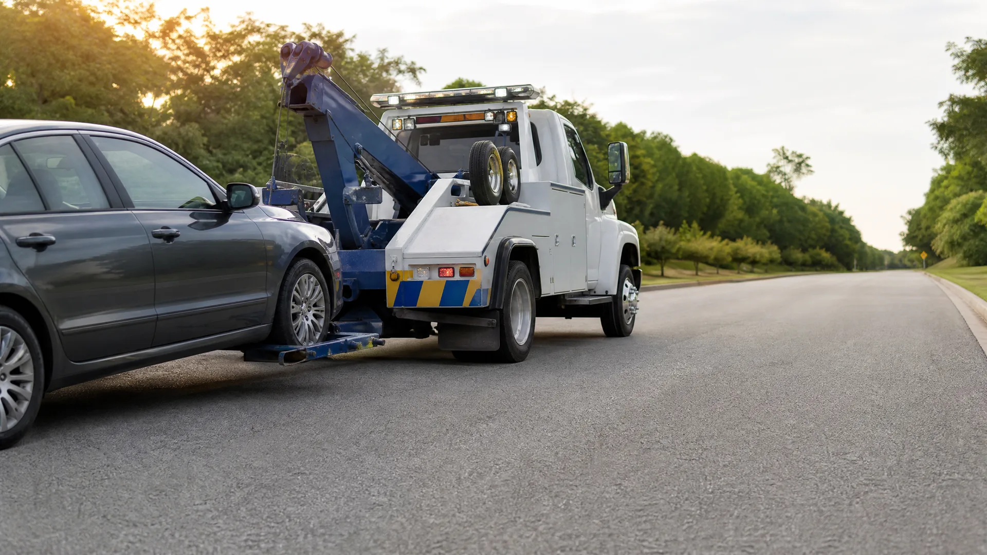 A white tow truck with blue and yellow chevron markings lifting the front of a gray sedan on a paved road lined with green trees.