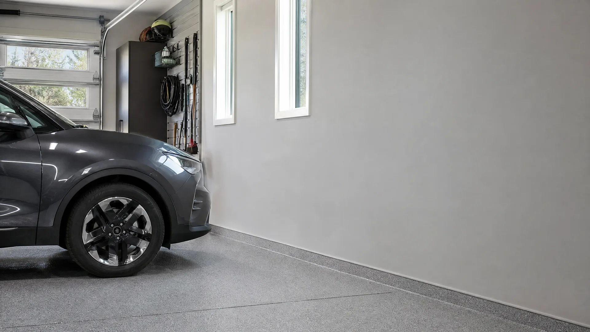 Interior of a garage with a gray flake-coated floor, light gray walls, two narrow windows, a dark storage cabinet with hanging tools, and the front half of a dark gray SUV parked beside a sectional garage door.