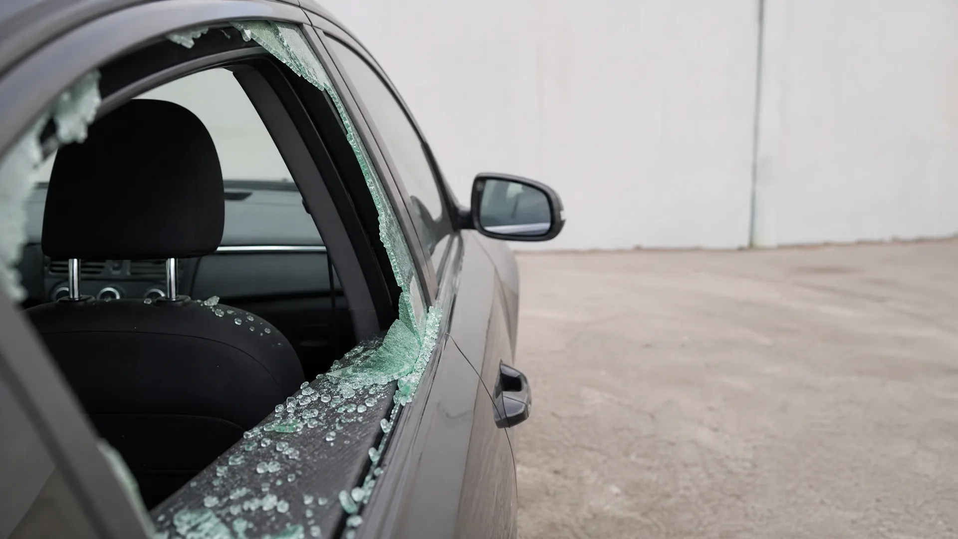 Close-up of a dark gray car with a shattered front door window, with glass fragments scattered on the door frame and seats, parked on bare ground beside a plain white wall.