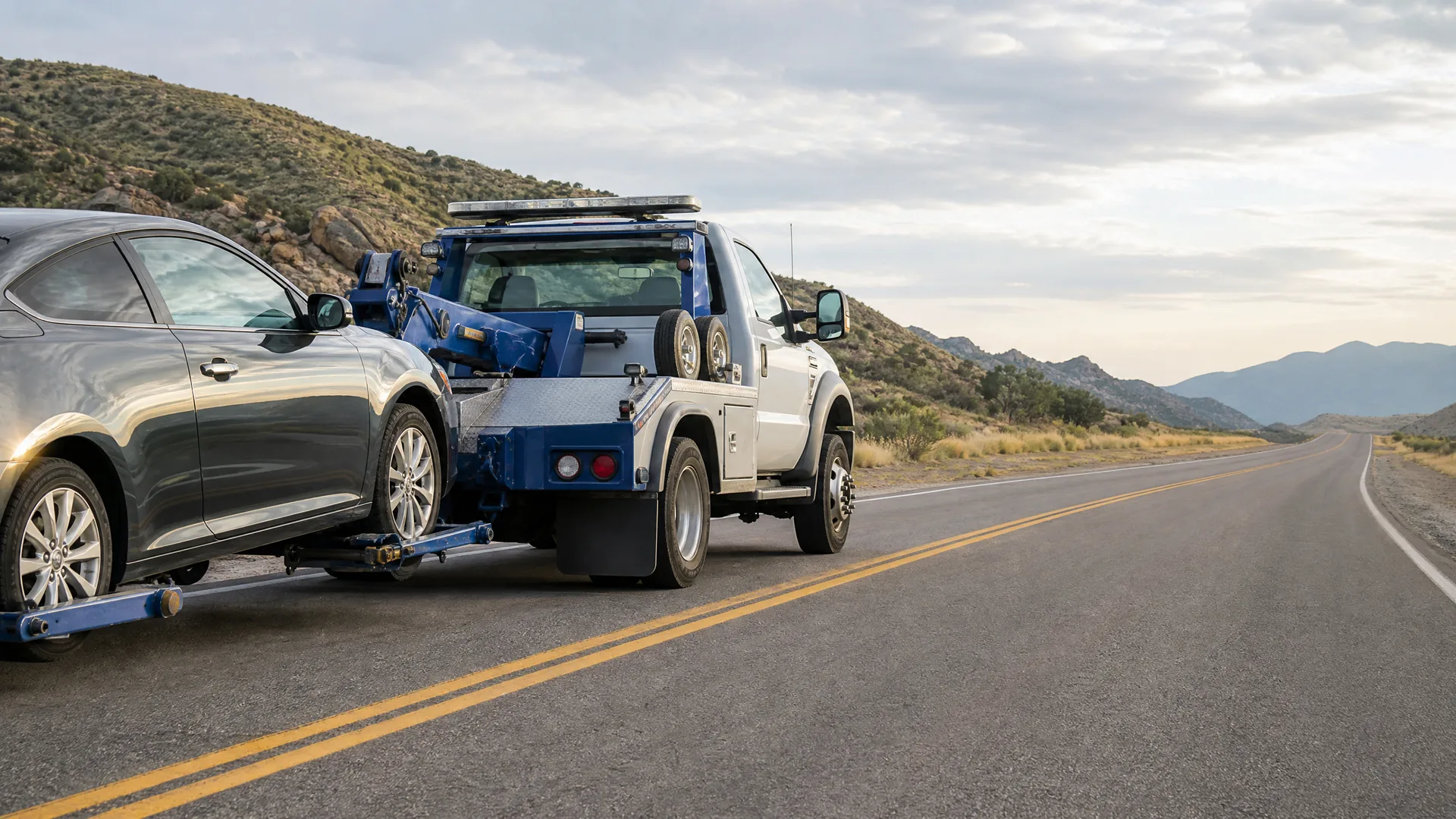 A white and blue tow truck parked on the shoulder of a two-lane road with yellow center lines, towing a dark gray coupe by its front wheels, with dry scrub hills and distant mountains in the background.