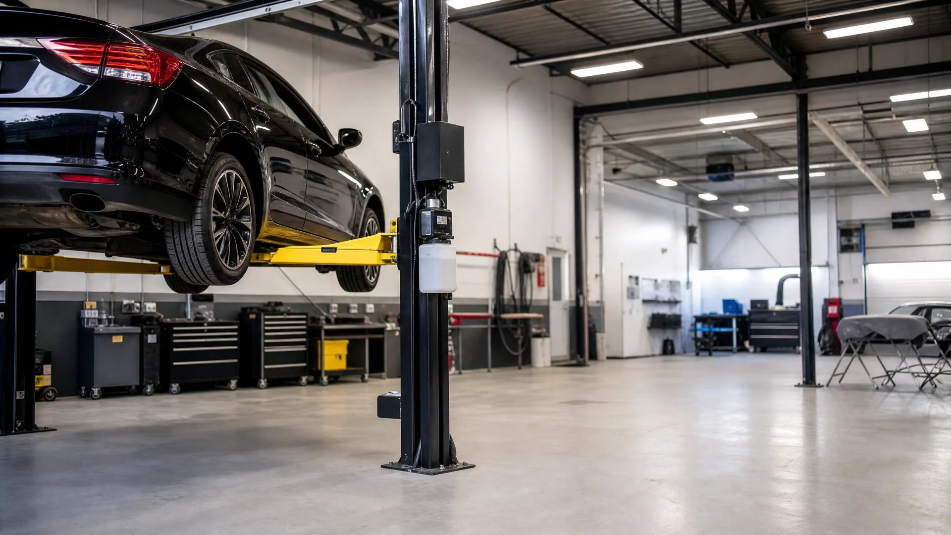 A black sedan raised on a yellow-armed two-post lift inside a workshop with tool cabinets, workbenches and overhead strip lighting.