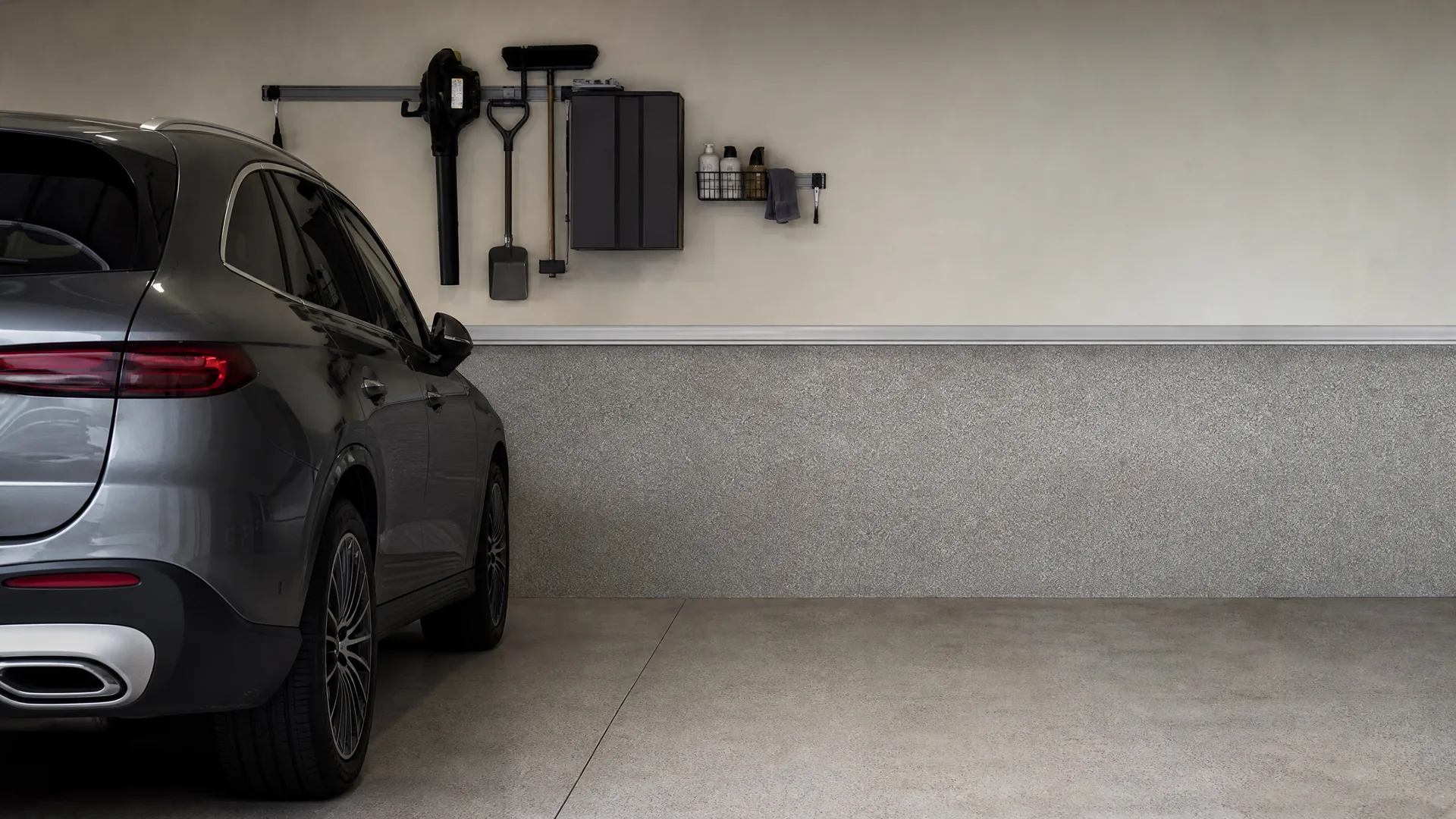 A gray SUV parked in a clean garage with a wall-mounted rack holding a leaf blower, shovel, broom and a cabinet, beside a beige wall with a speckled lower panel.