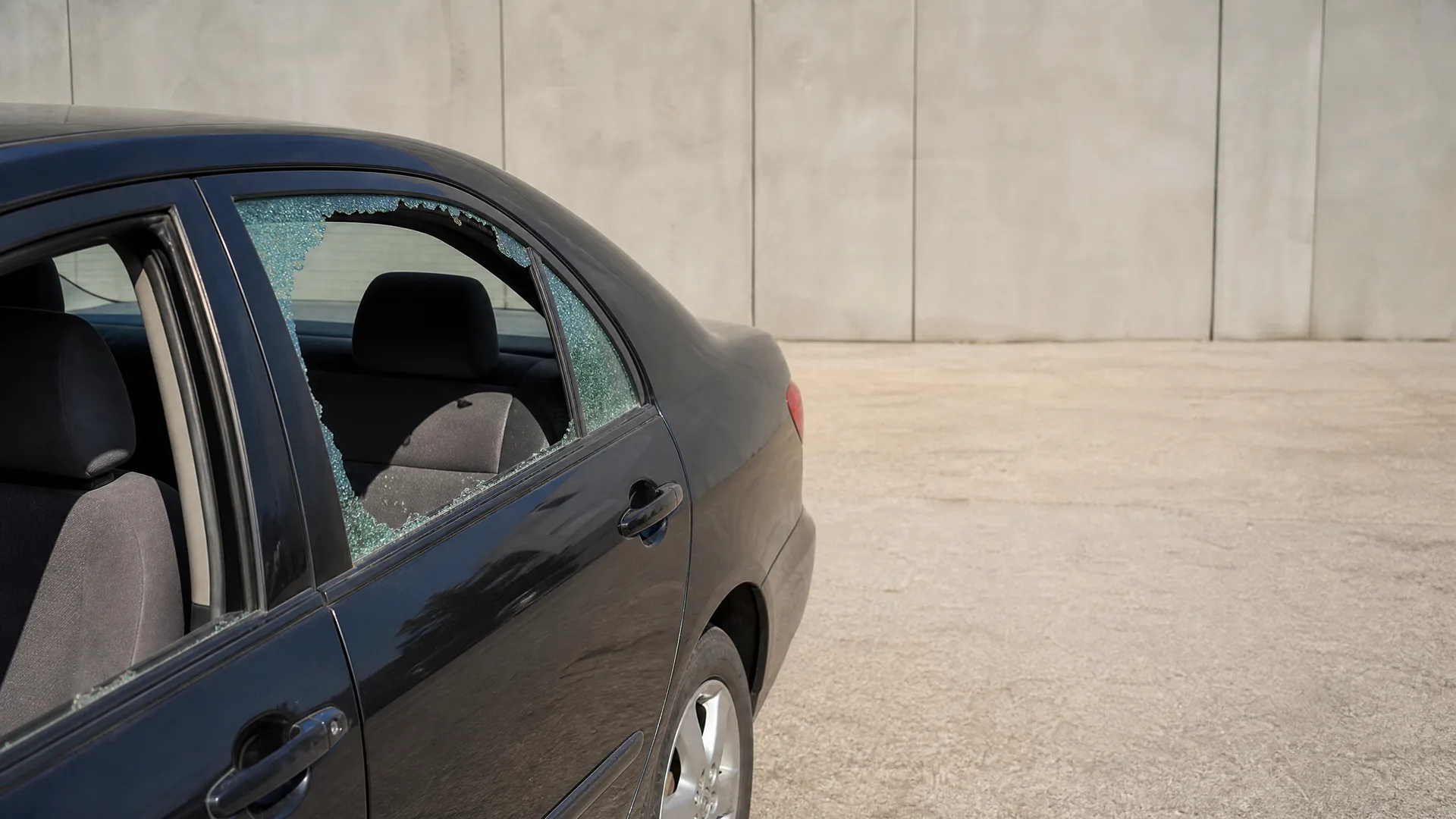 A dark blue sedan parked on pavement beside a plain concrete panel wall, with its rear passenger window shattered and broken glass fragments along the frame.
