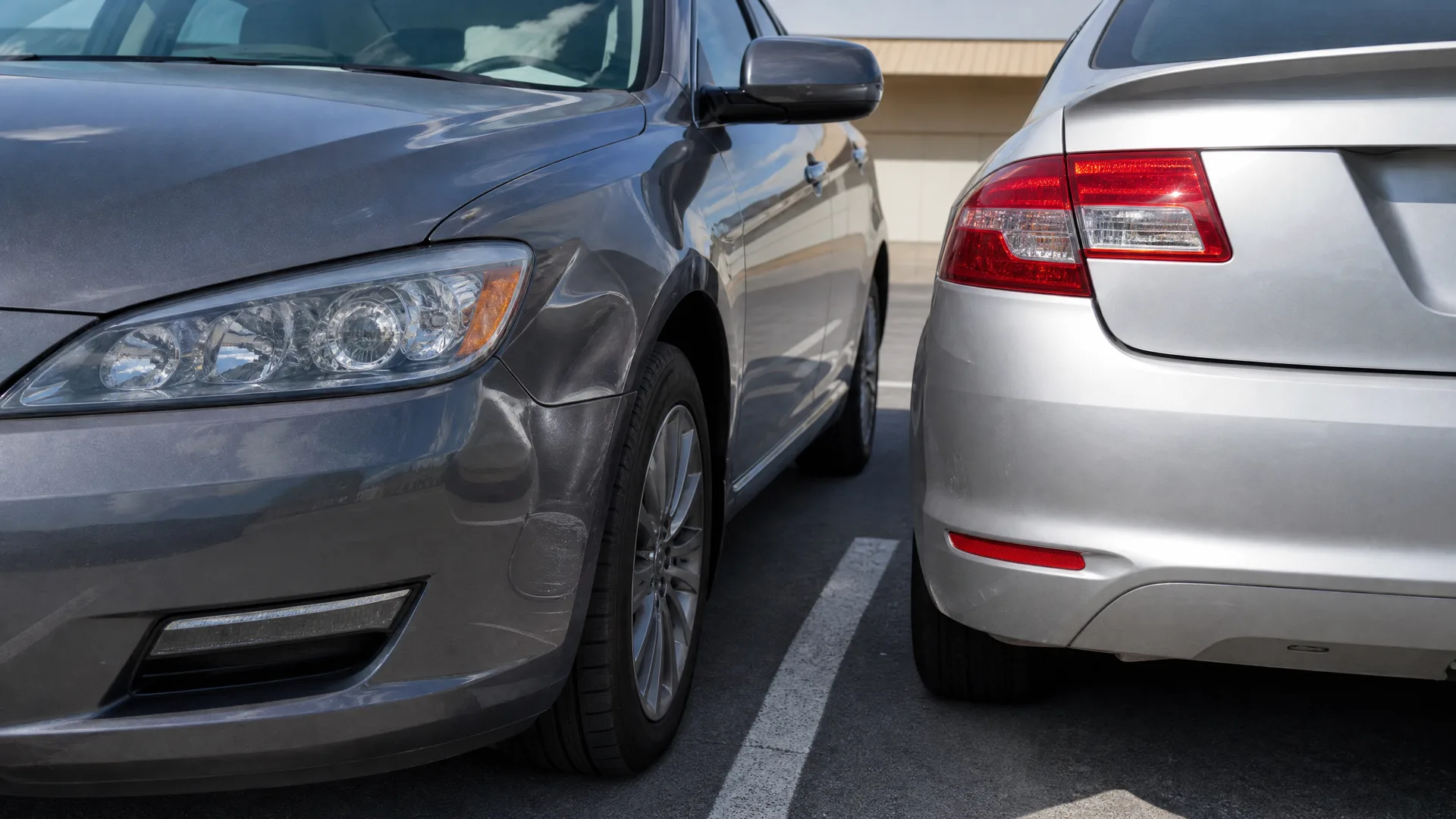 Close-up of two parked cars nearly touching, a gray sedan's front end on the left and a silver sedan's rear end on the right, with a painted parking line on the asphalt between them.