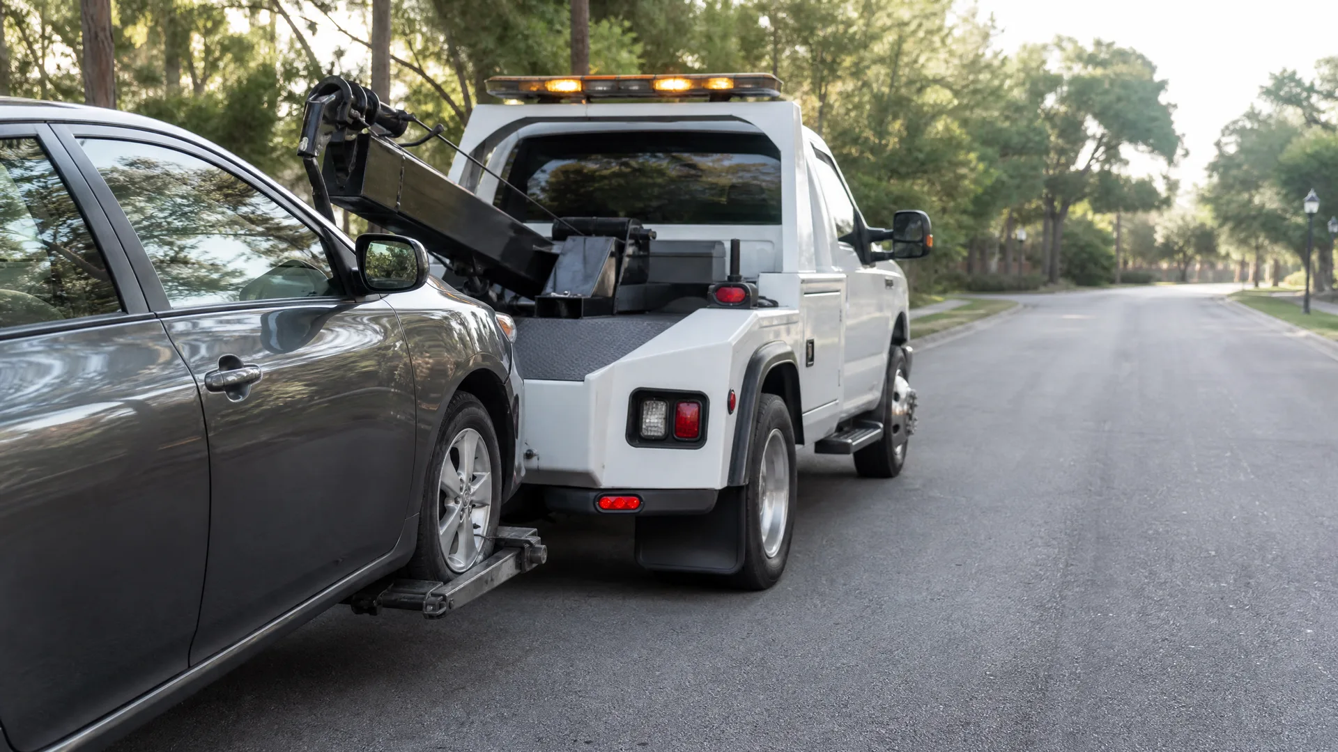 A white tow truck with a wheel-lift towing a gray sedan by its front wheels on a paved residential street lined with trees.
