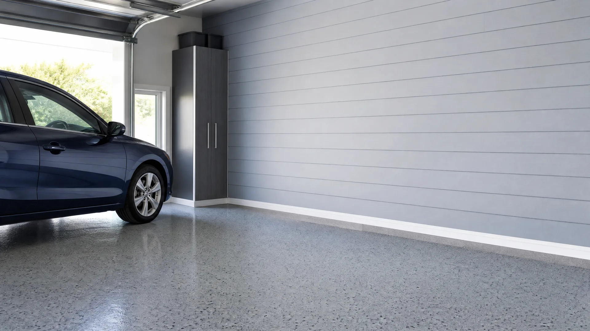 Interior of a garage with a speckled epoxy floor, light gray horizontal plank walls, a tall dark storage cabinet in the corner, and the front half of a dark blue car parked by the open garage door.