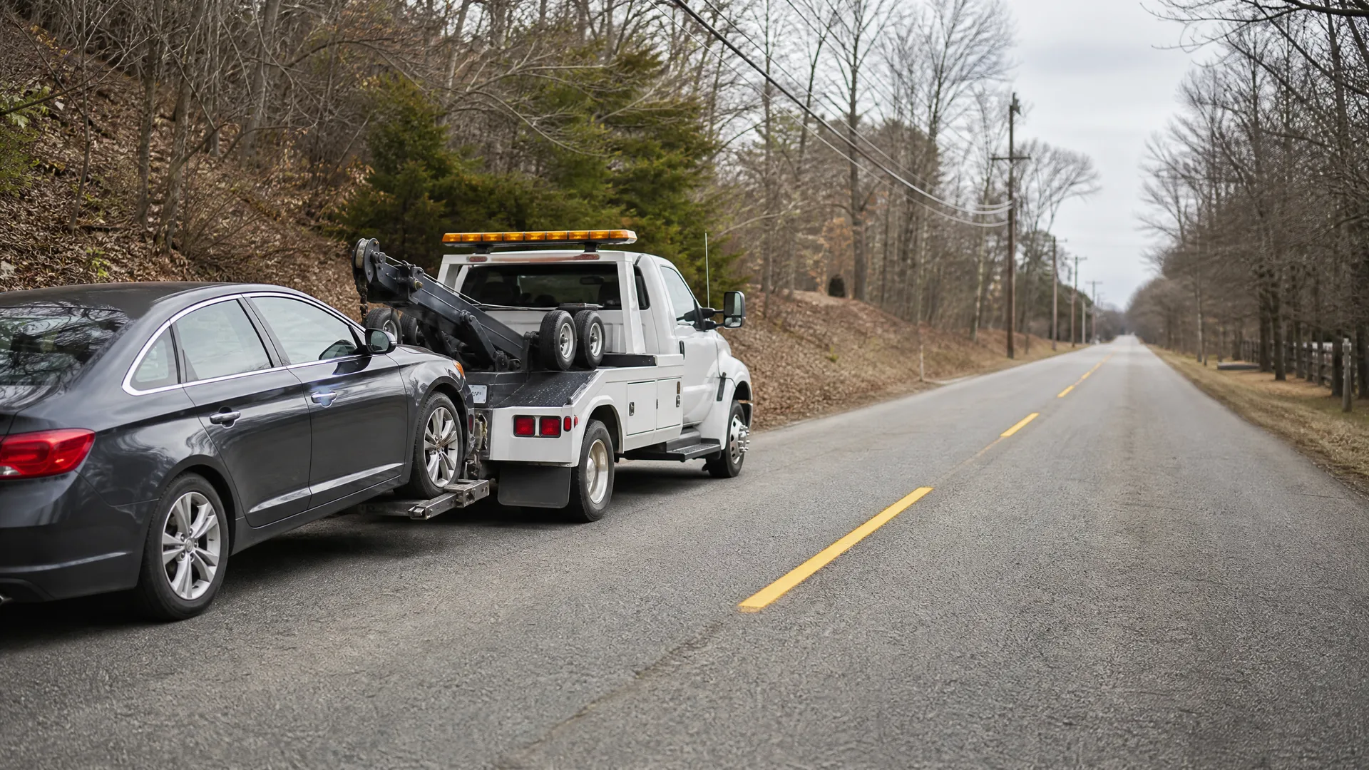 A white flatbed tow truck parked on the shoulder of a two-lane road with a dark gray sedan loaded on its bed, with leafless trees and a wooded slope alongside.