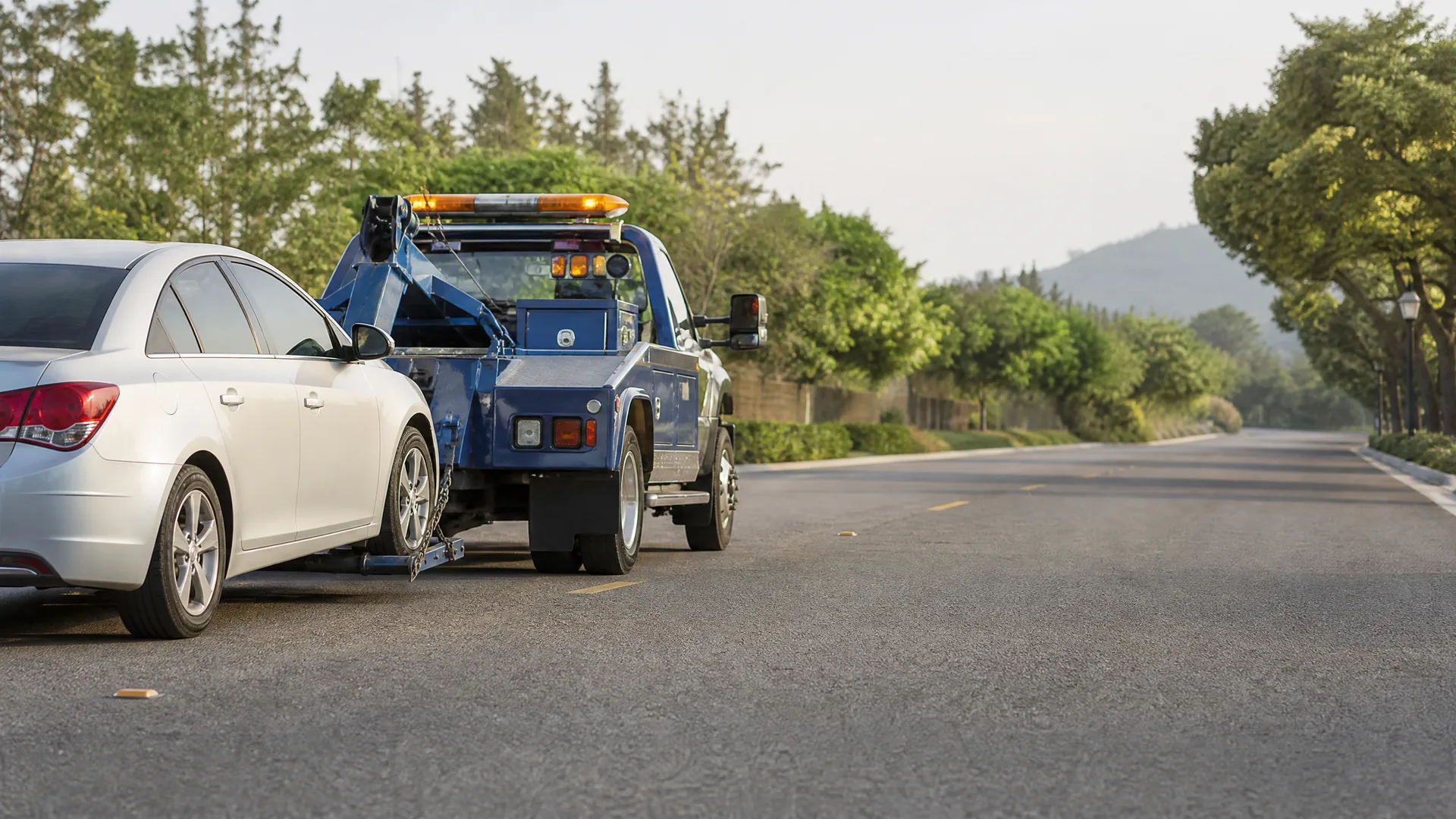 A blue tow truck tows a silver sedan by its front wheels along a paved road lined with green trees, with a low hill in the background.