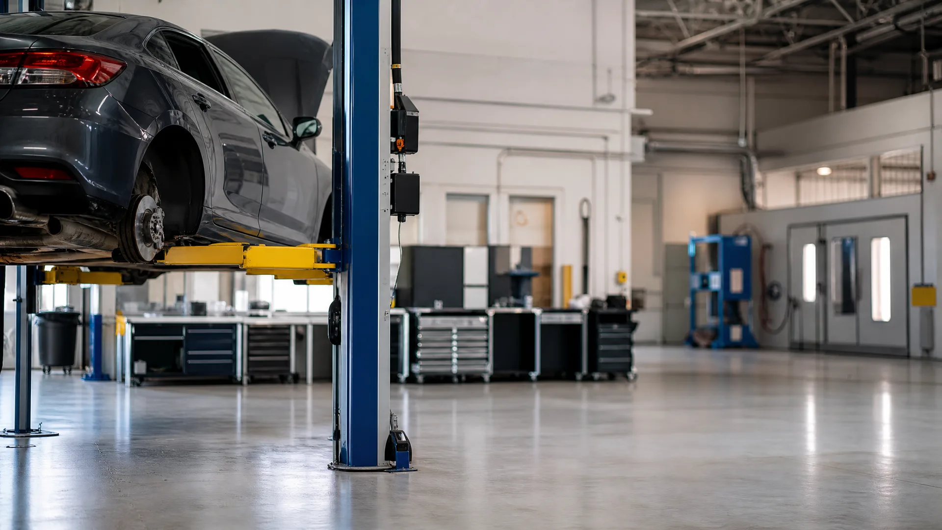 Interior of an auto repair shop with a gray sedan raised on a blue two-post lift, tool cabinets and workbenches along the wall, and a polished concrete floor.