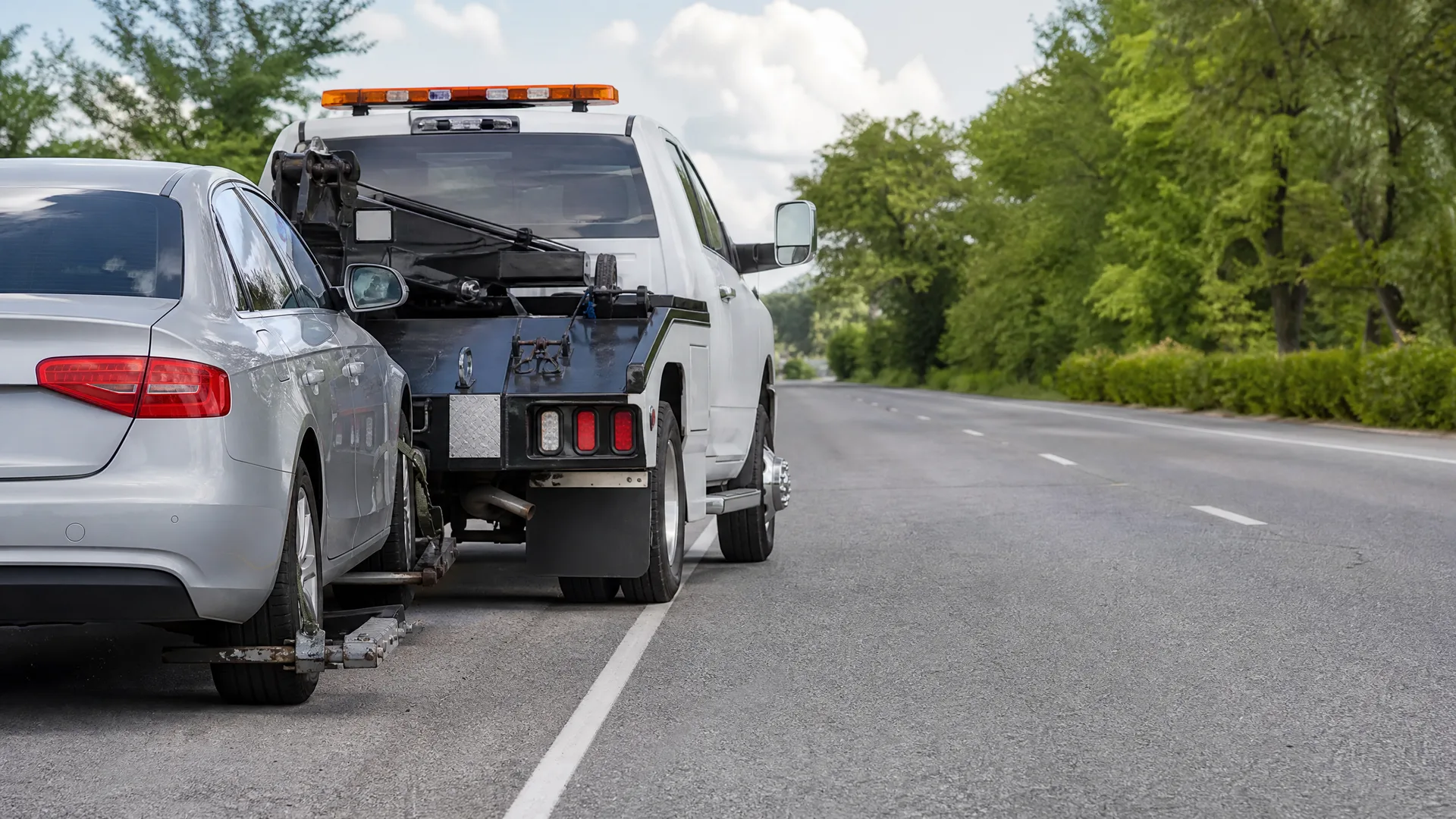 A white tow truck with an orange light bar towing a silver sedan by its front wheels at the edge of an asphalt road lined with green trees.