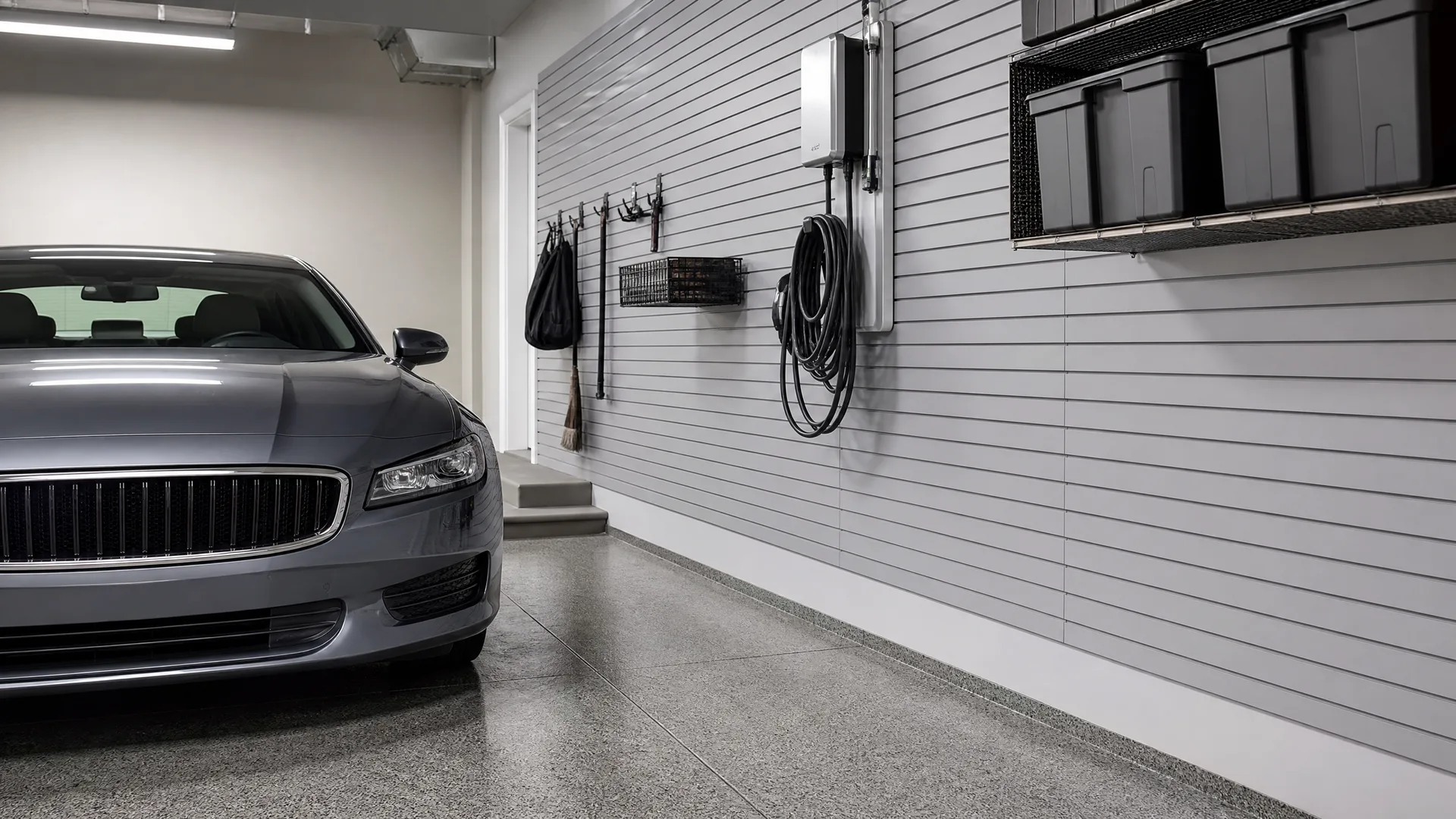 Interior of a garage with a polished concrete floor, a gray car parked at left, and a gray slatwall on the right holding a wall-mounted unit with a coiled cable, hanging baskets, a broom, and overhead shelving with black bins.