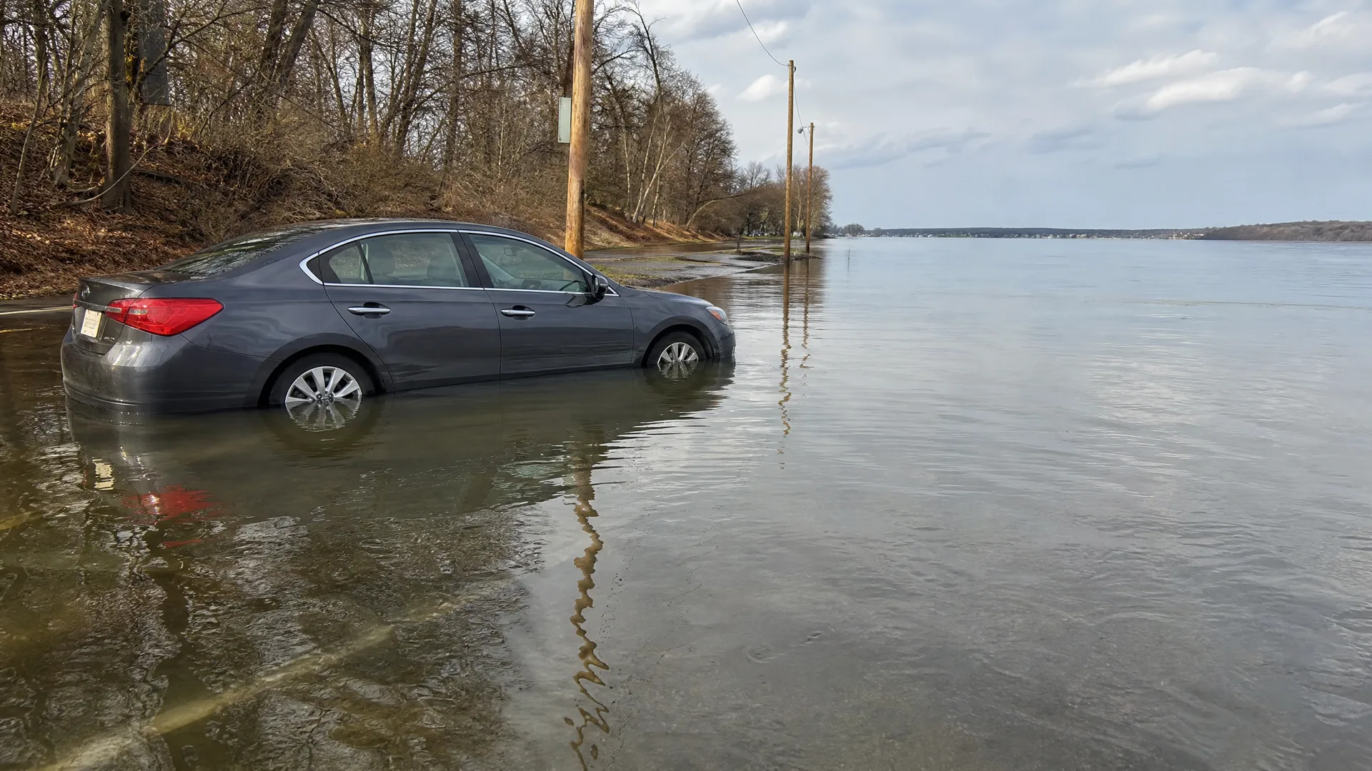 A gray sedan sits in floodwater covering a road beside a wooded bank, with a wide expanse of water and utility poles extending to the horizon under a partly cloudy sky.