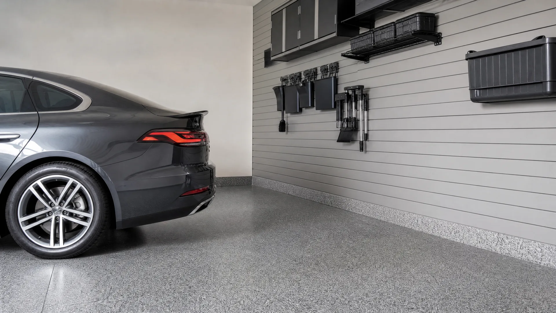 Garage interior with a speckled epoxy floor, a dark gray sedan parked at left, and a light gray slatwall holding black cabinets, wall-mounted bins, shelves, and hanging tools.