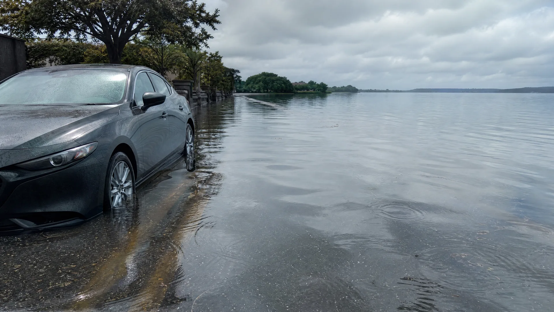 A dark sedan parked at the edge of a flooded road, with water covering the street and extending to a distant tree-lined shore under an overcast sky.
