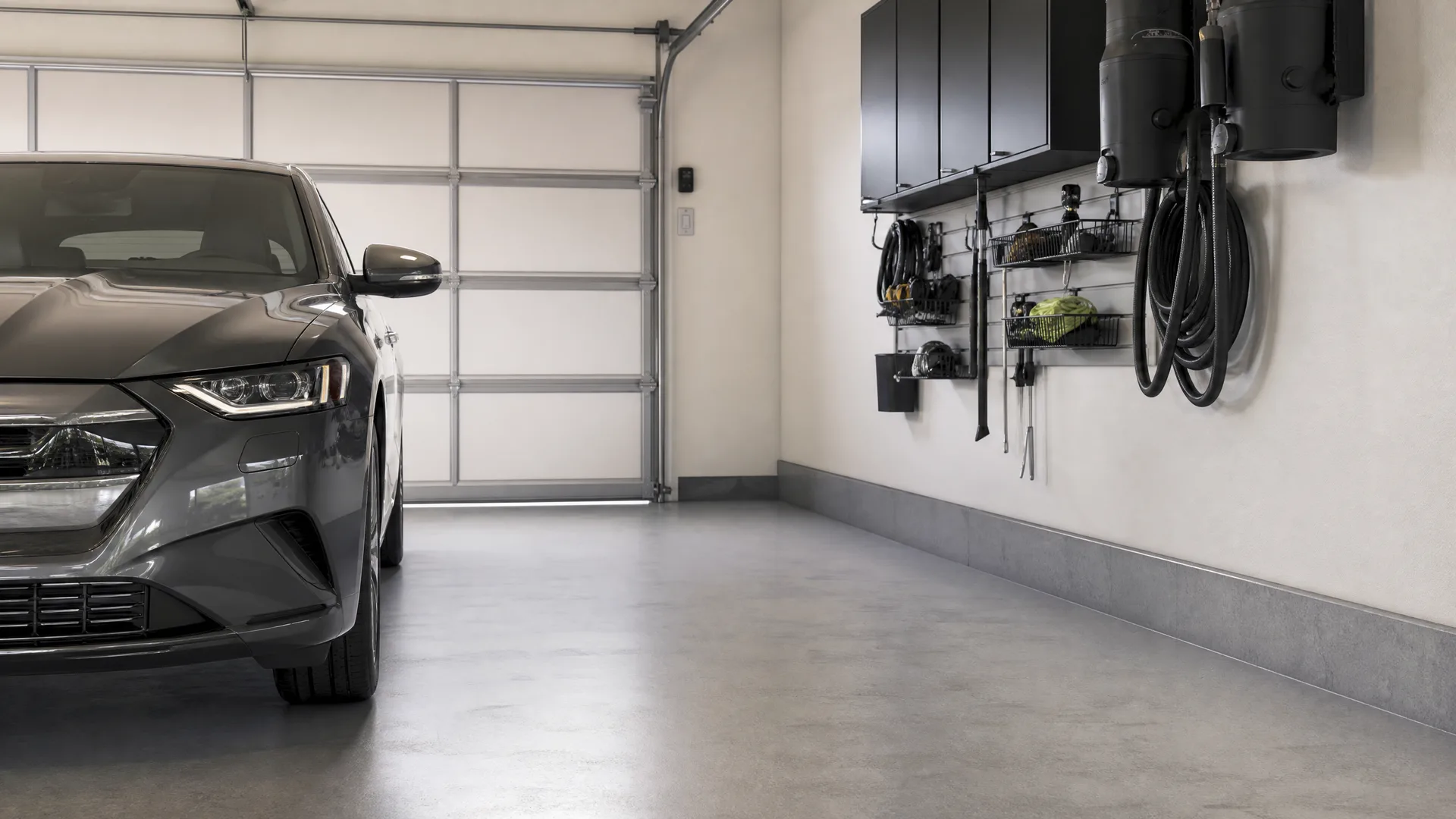 Interior of a garage with a gray car parked at left beside a white sectional door, a polished concrete floor, and a wall at right with black overhead cabinets, a tool rack, and coiled hoses.