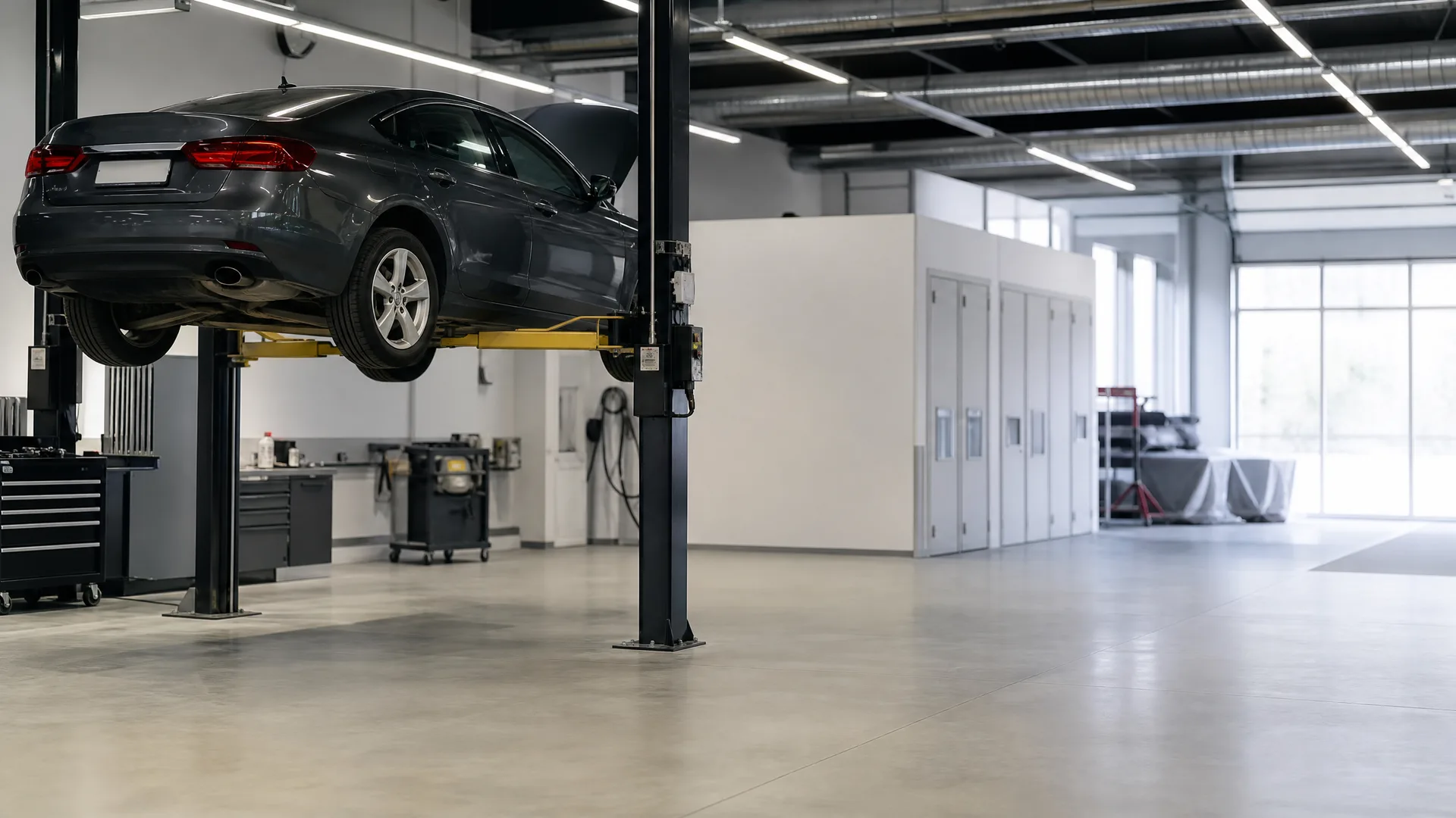 A dark grey sedan raised on a two-post lift inside a bright, empty vehicle workshop with polished concrete floors, tool cabinets, and white partition walls.