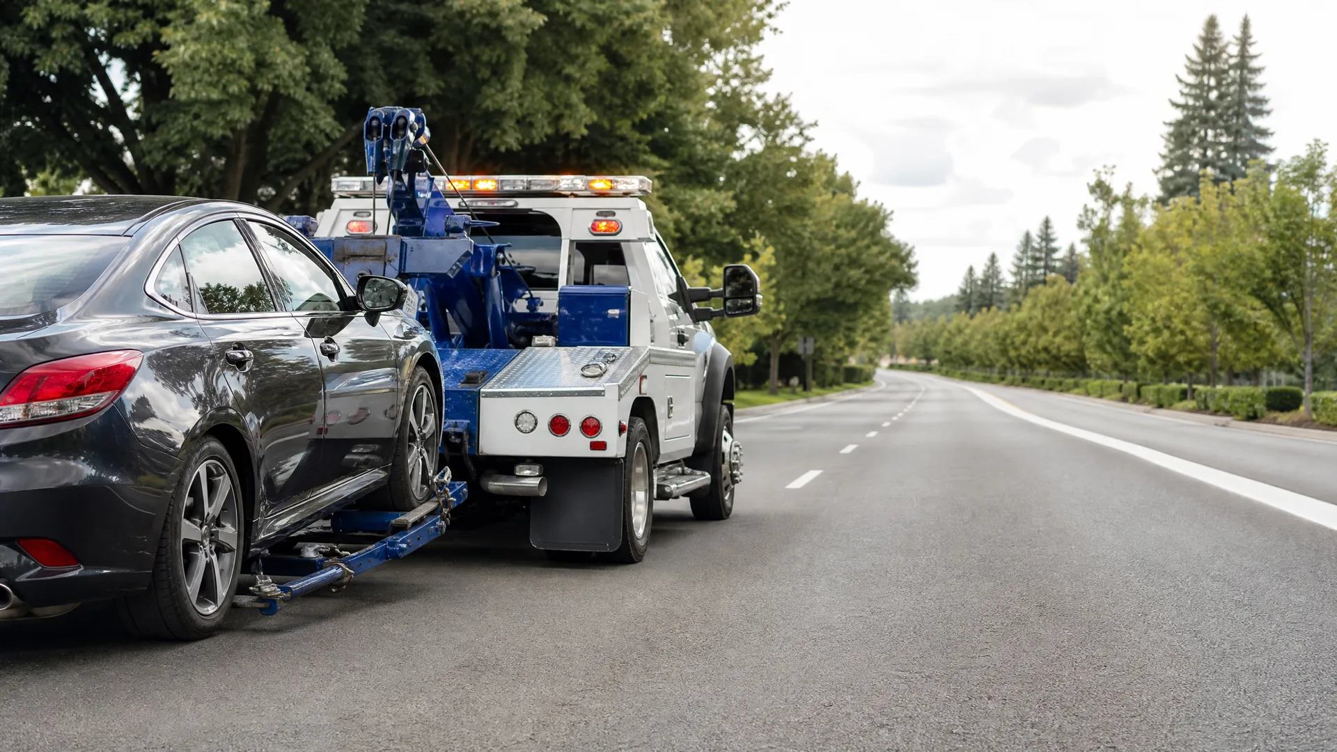 A blue and white tow truck parked on a road shoulder towing a dark gray sedan by its front wheels, with trees lining both sides of the empty road.