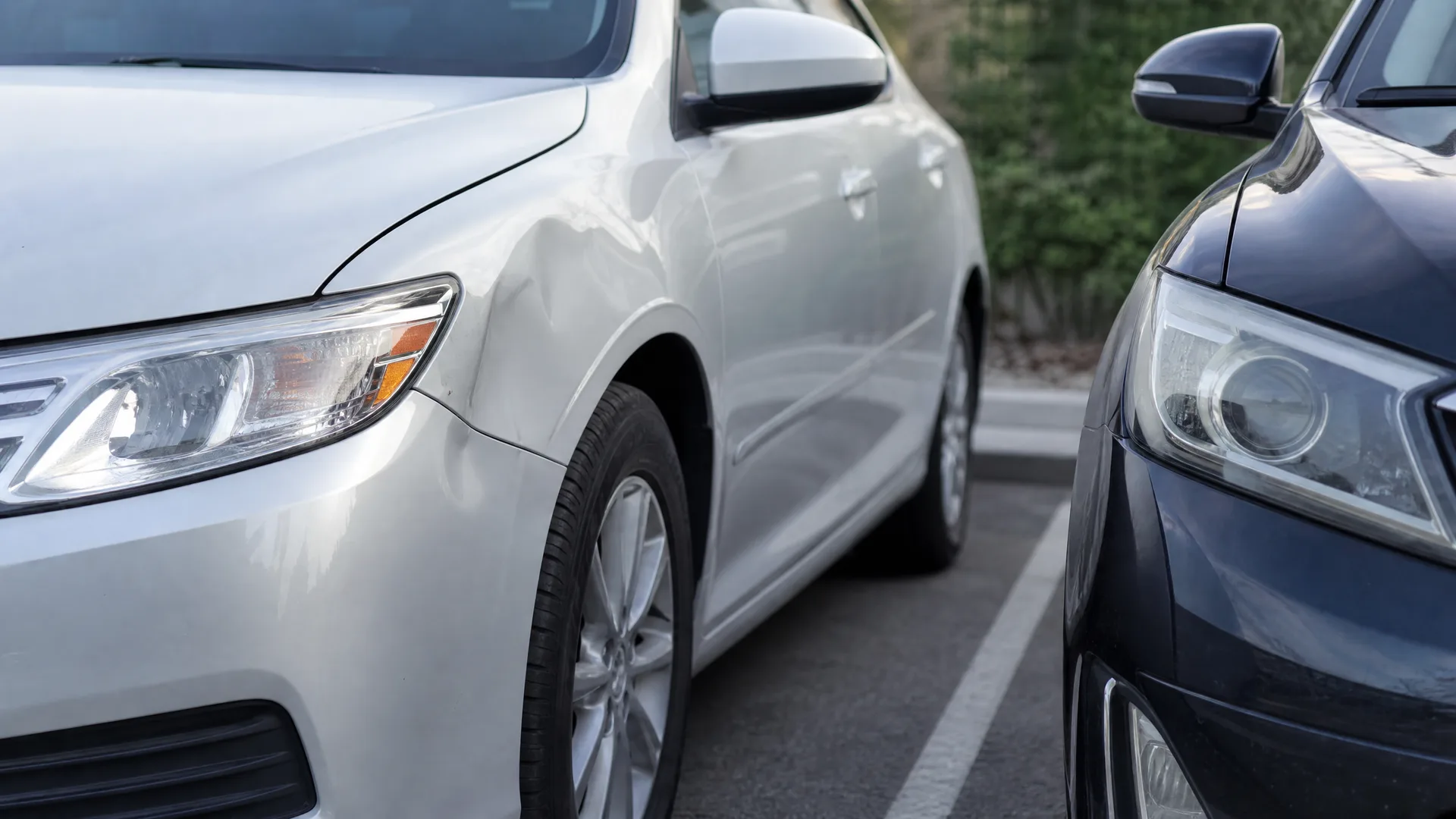 Close-up of a silver sedan with a dent in its front fender parked beside a dark gray car in a marked parking lot, with green shrubs in the background.