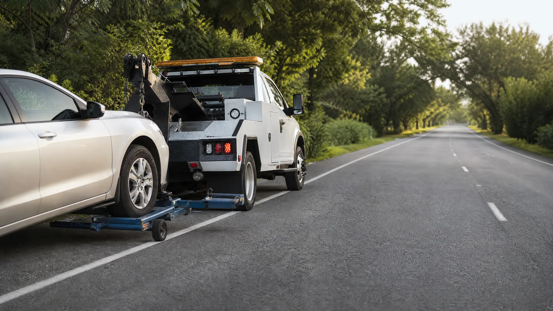 A white tow truck with a wheel-lift dolly towing a silver car, parked on the shoulder of a tree-lined two-lane road.