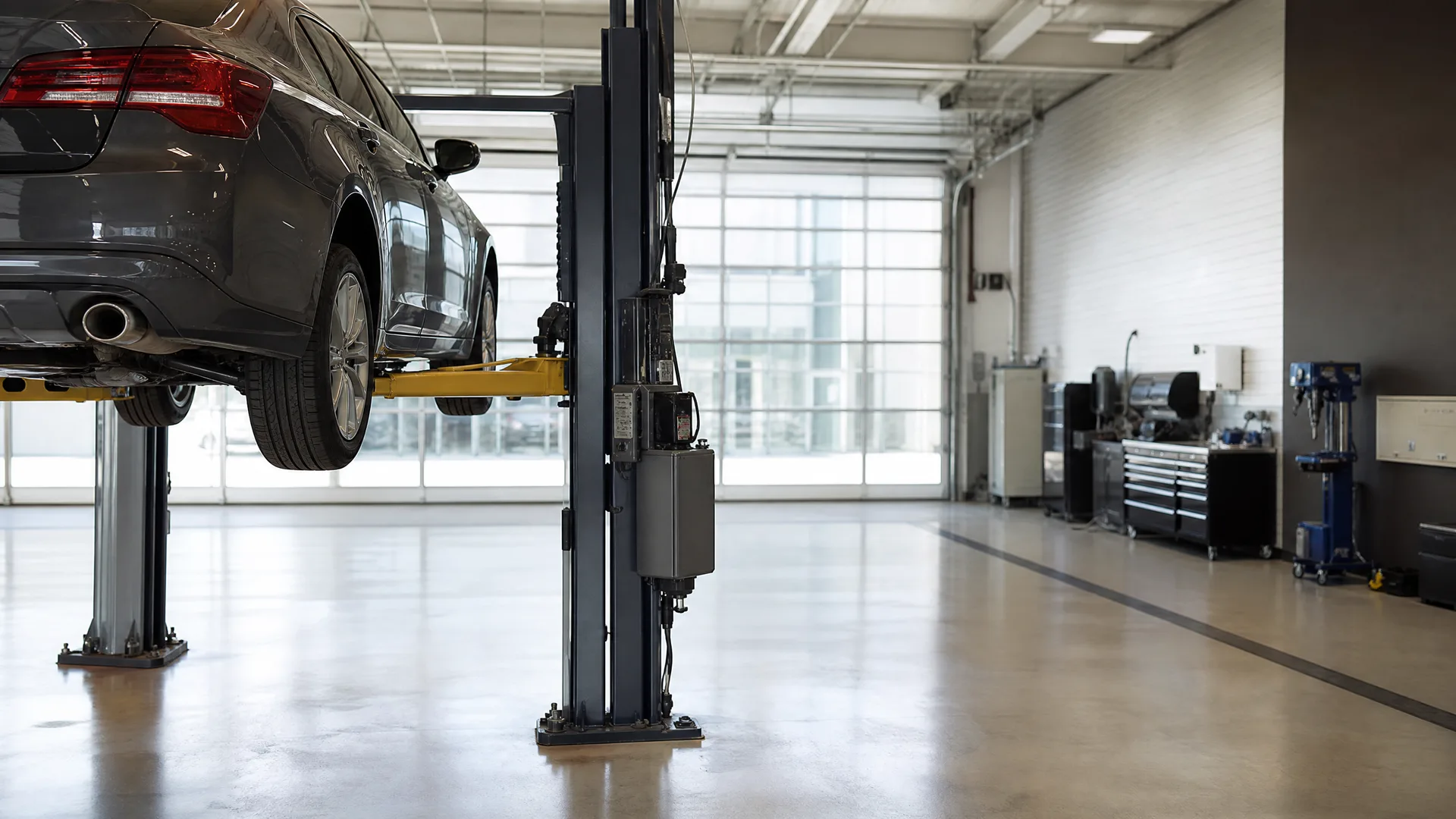 A dark gray sedan raised on a two-post hydraulic lift inside a service garage with polished concrete floors, large windows, tool cabinets and equipment along the right wall.